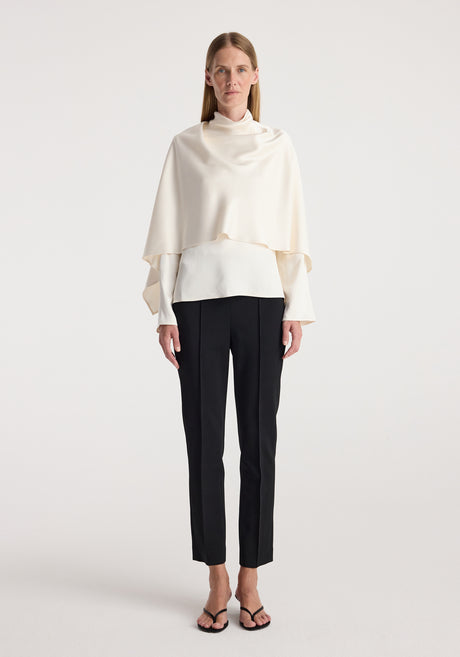 Woman wearing a cream satin cape top with long sleeves paired with black tailored pants and open-toe heels standing against a plain white background.