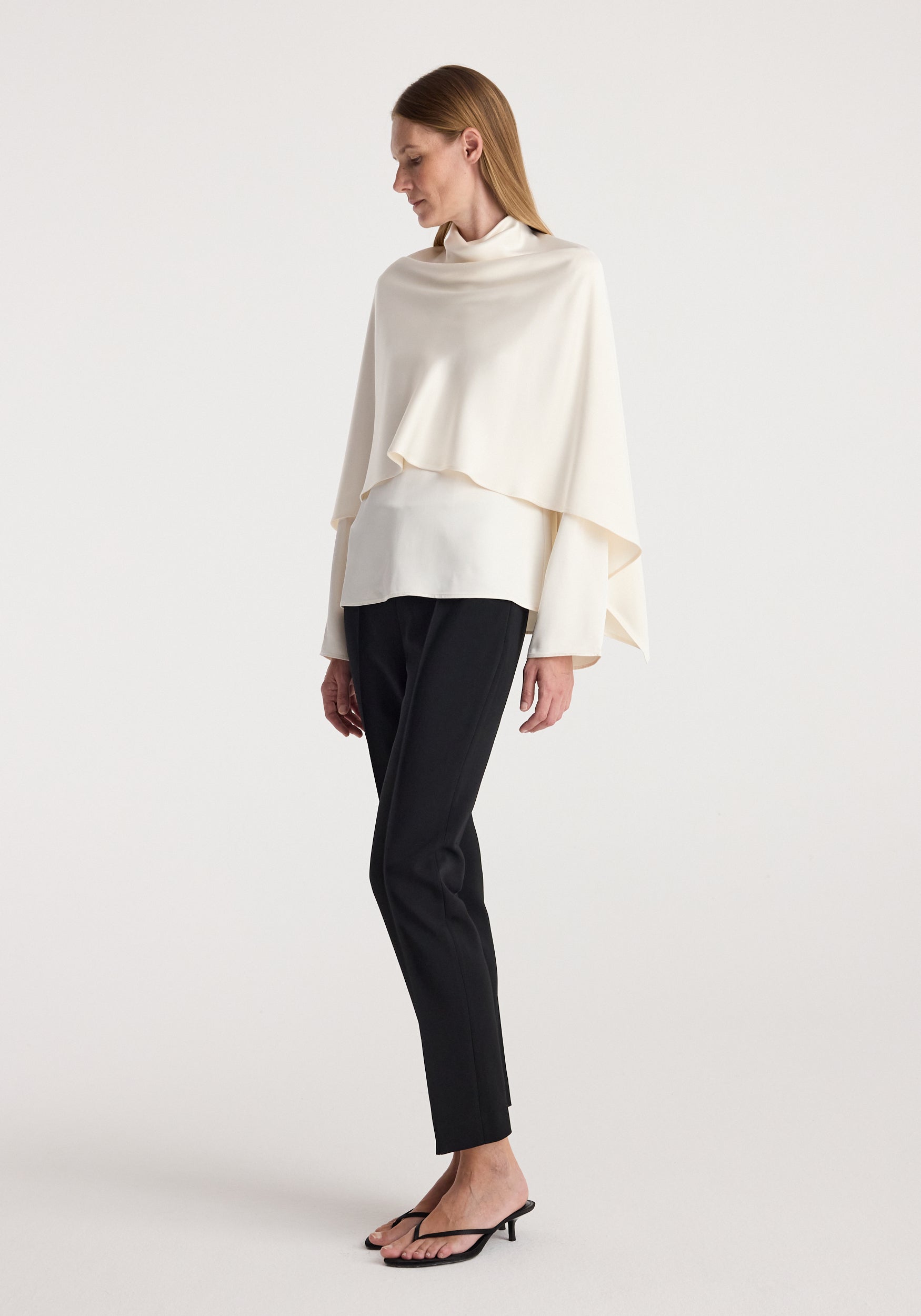 Woman wearing a cream draped satin cape top over a long-sleeve shirt, paired with black fitted trousers and black heeled sandals, standing against a plain background in satin cape top | cream.