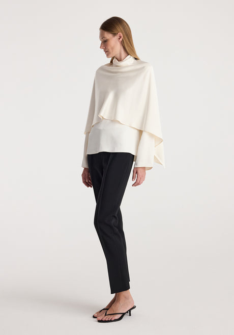 Woman wearing a cream satin cape top with long sleeves paired with black tailored pants and open-toe heels standing against a plain white background.