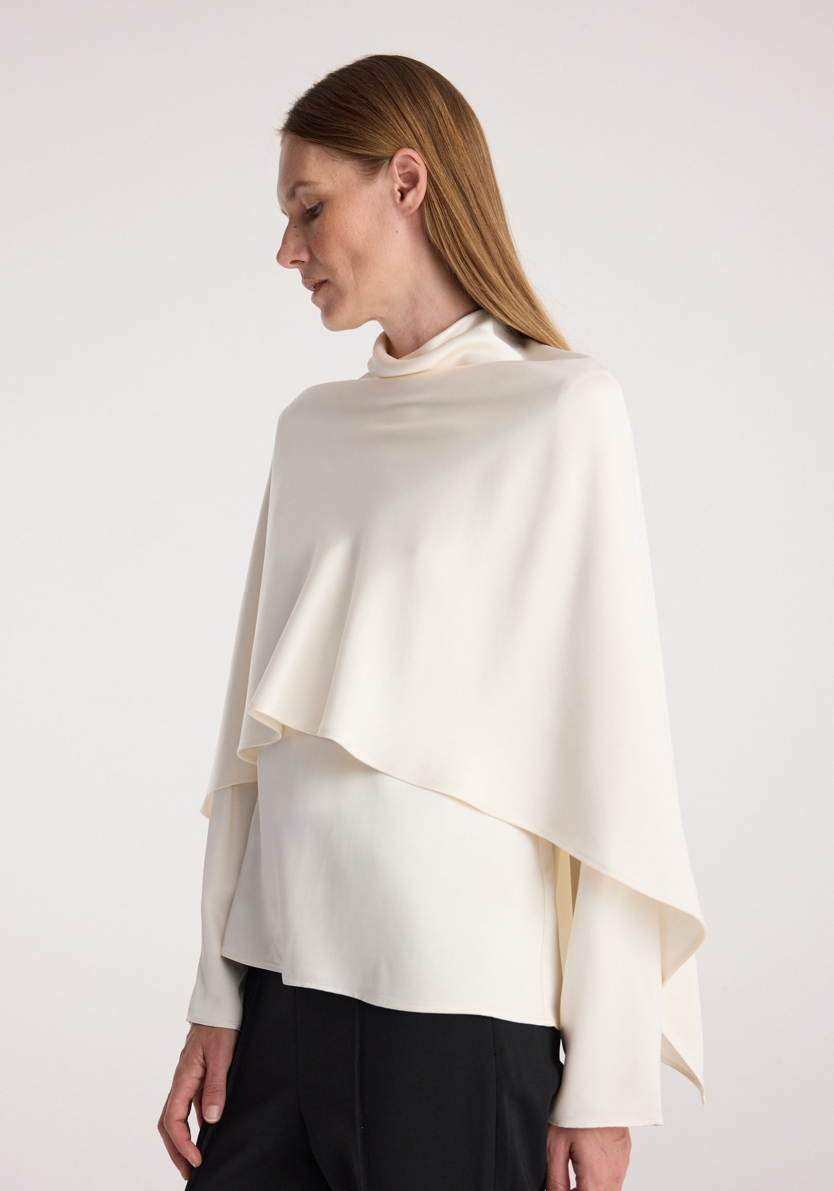 Woman wearing a cream-colored Satin cape top | cream with a high neckline and draped asymmetrical cape detail over a fitted long sleeve shirt paired with black pants.