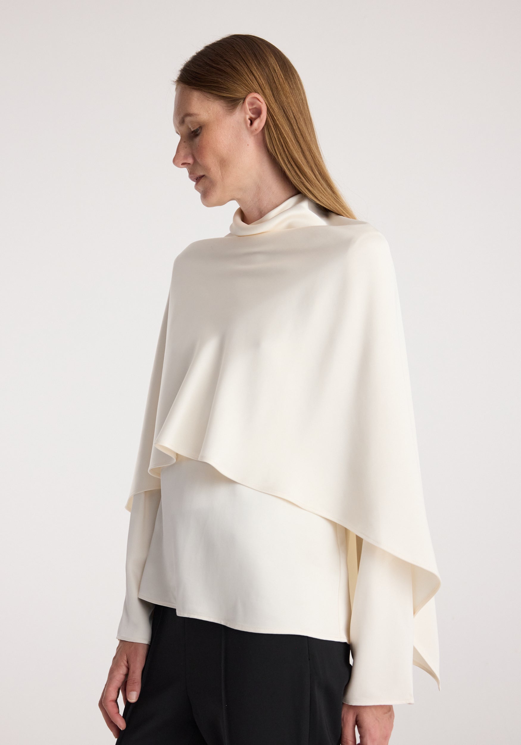 Woman wearing a cream-colored Satin cape top | cream with a high neckline and draped asymmetrical cape detail over a fitted long sleeve shirt paired with black pants.