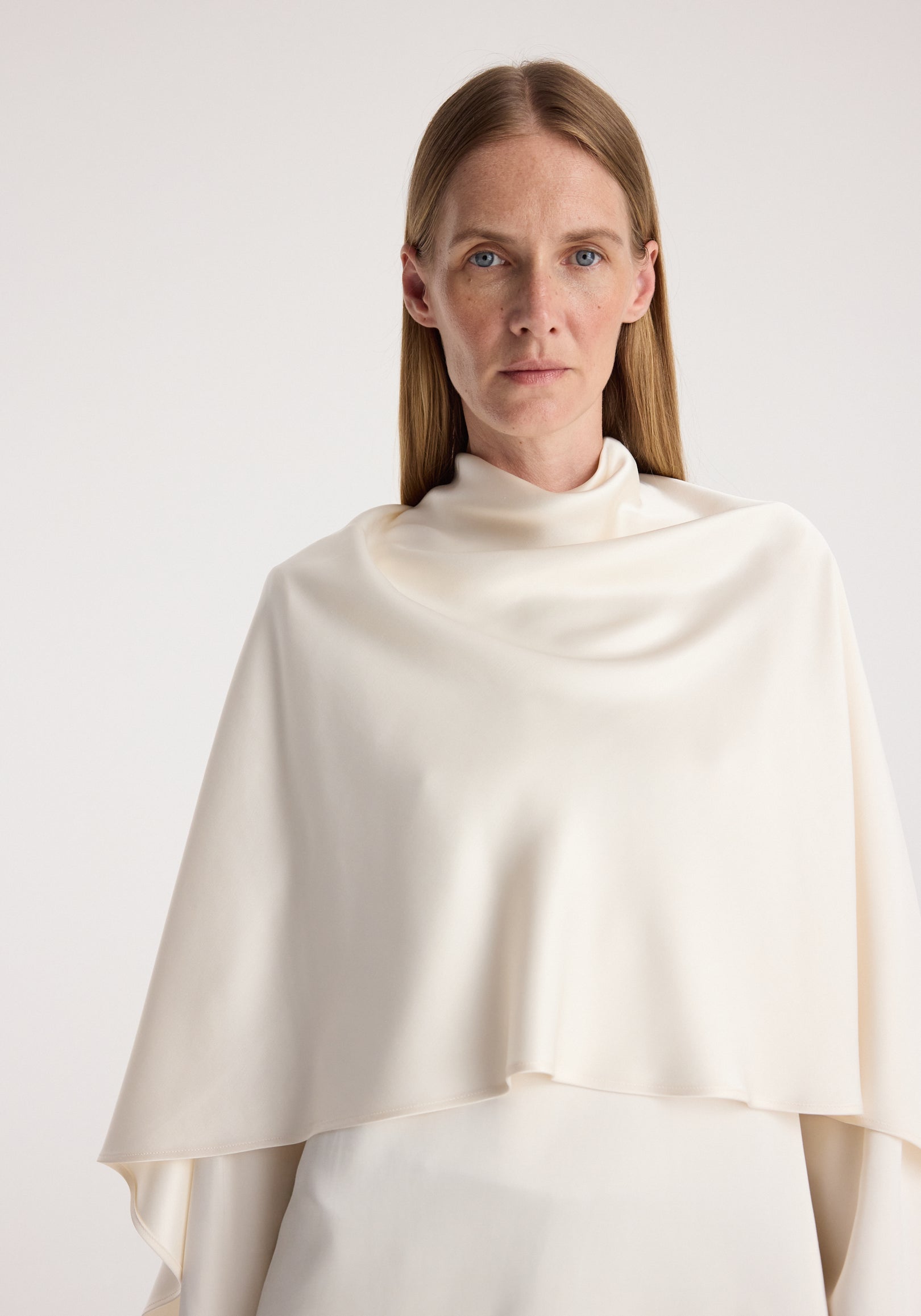 Woman wearing a cream satin cape top | cream with a smooth, draped design and high neckline, posing against a plain light background.