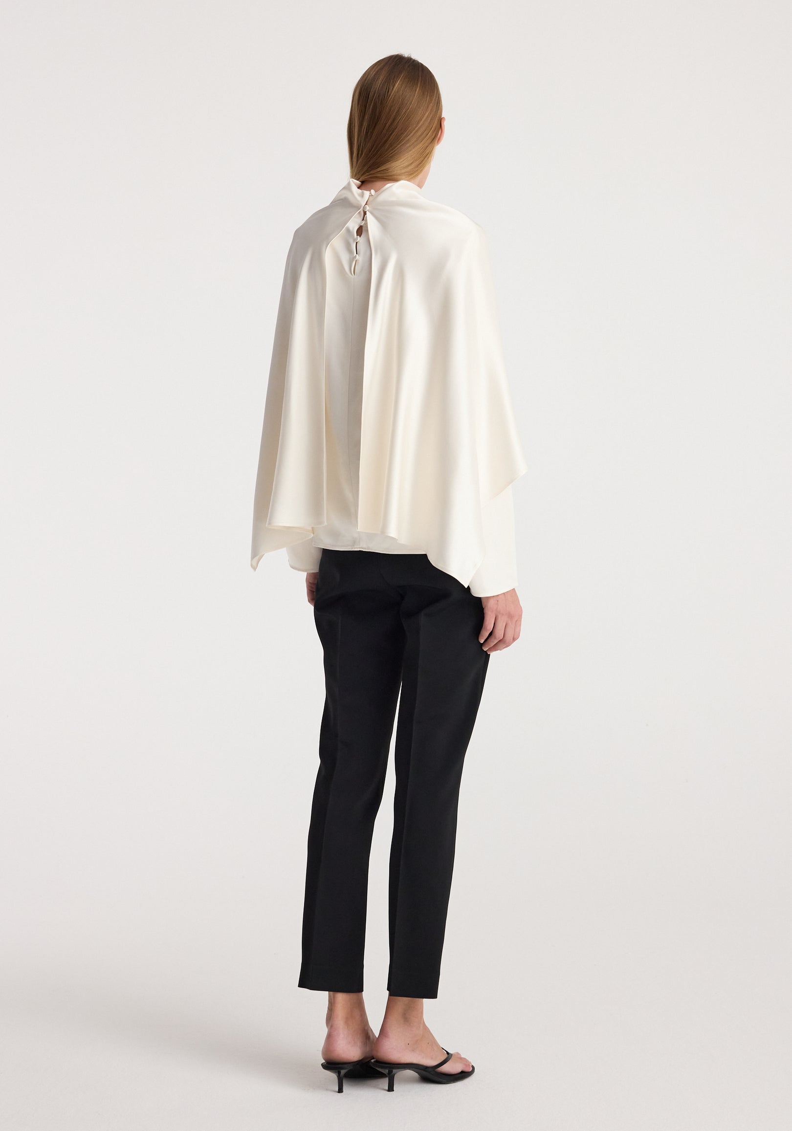 Rear view of a model wearing a flowing satin cape top | cream paired with black slim pants and black heeled sandals, shown against a plain white background.