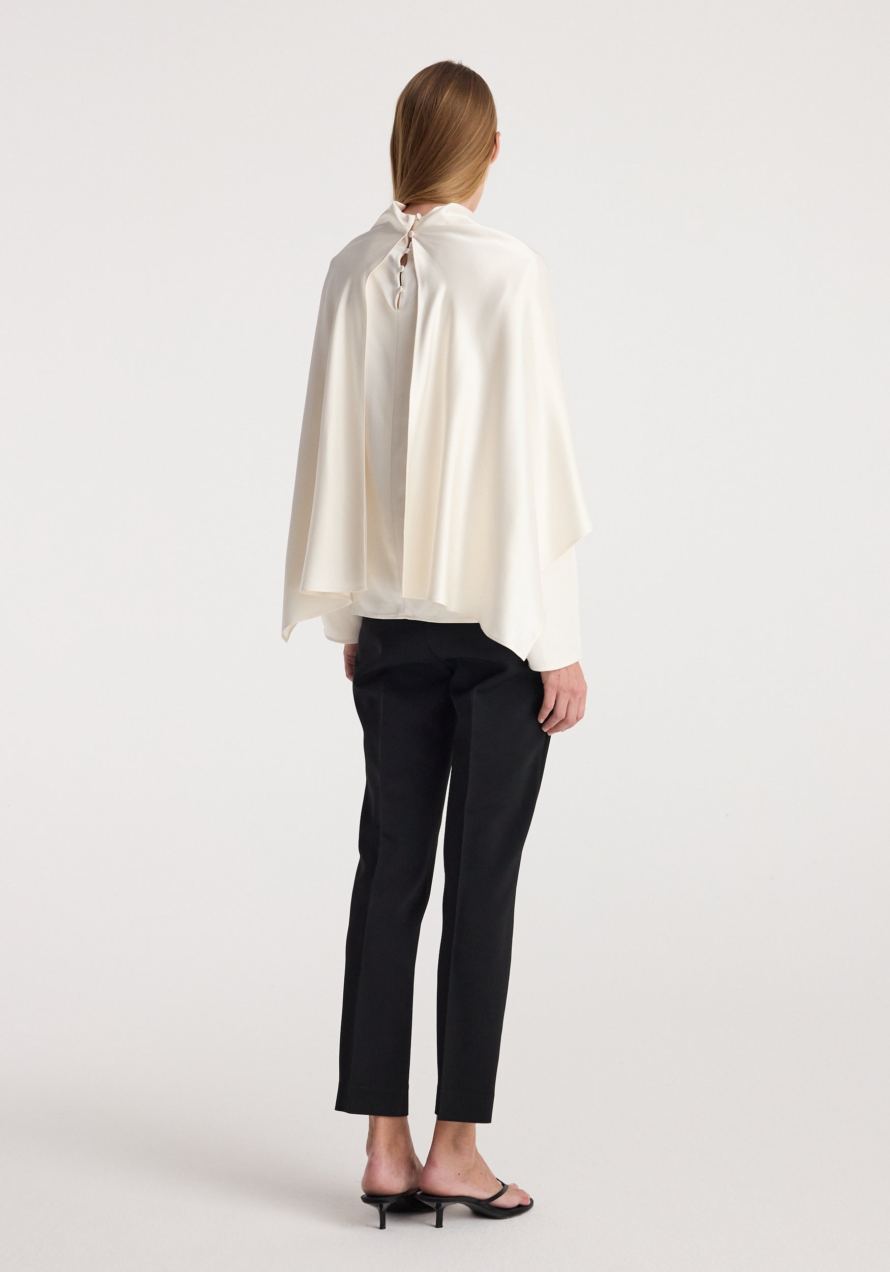 Rear view of a model wearing a flowing satin cape top | cream paired with black slim pants and black heeled sandals, shown against a plain white background.