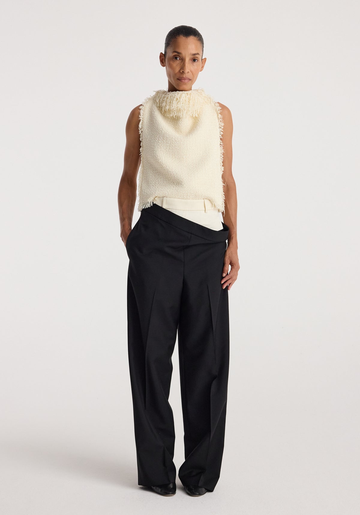 Model wearing a sleeveless textured fringe bouclé top | cream paired with loose black trousers, standing against a plain white background.