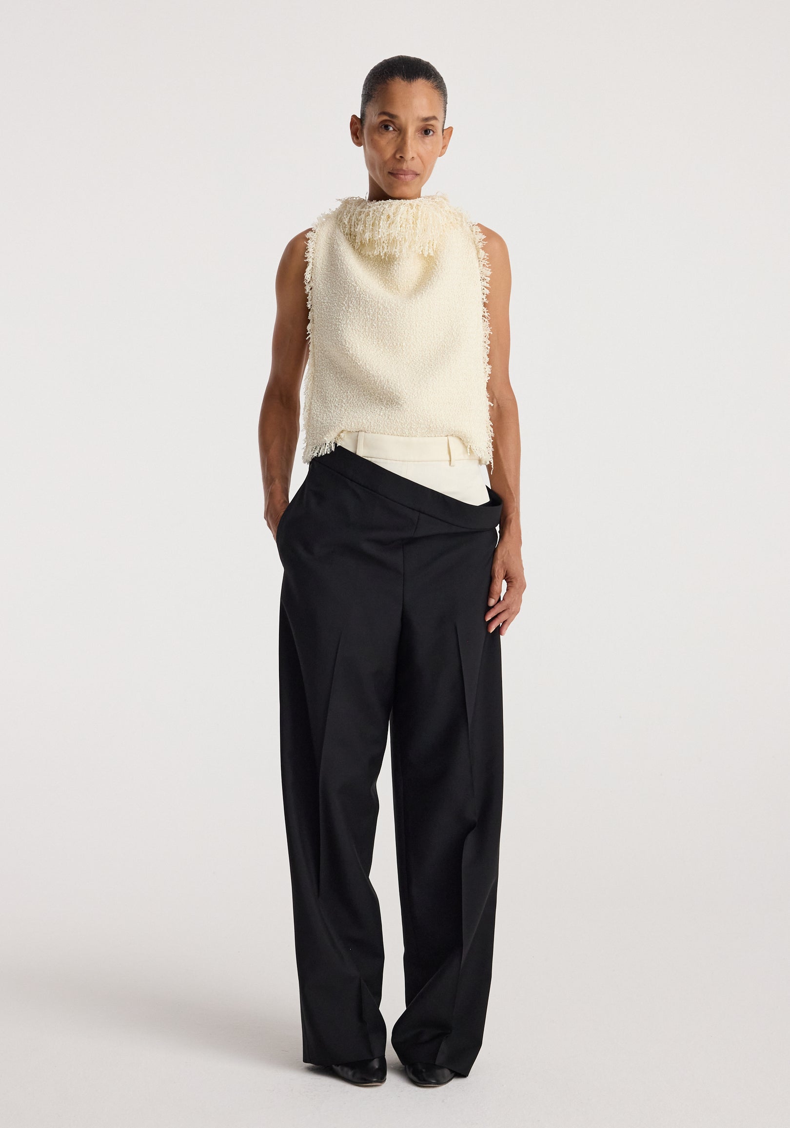 Model wearing a sleeveless textured fringe bouclé top | cream paired with loose black trousers, standing against a plain white background.