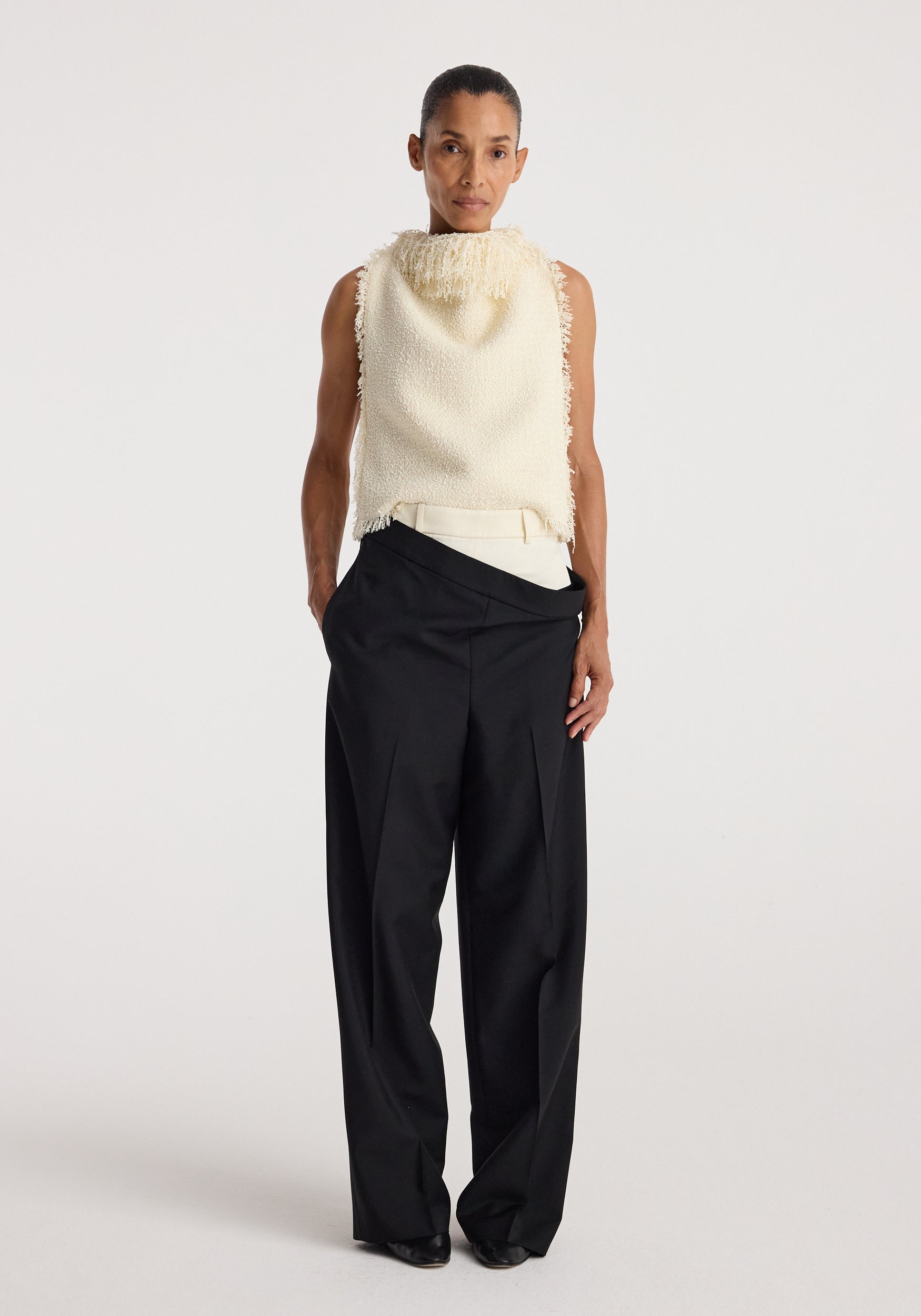 Model wearing a sleeveless textured fringe bouclé top | cream paired with loose black trousers, standing against a plain white background.