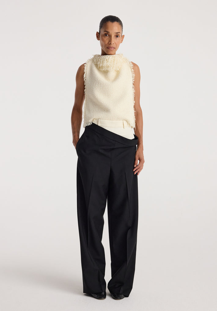 Model wearing a sleeveless textured fringe bouclé top | cream paired with loose black trousers, standing against a plain white background.