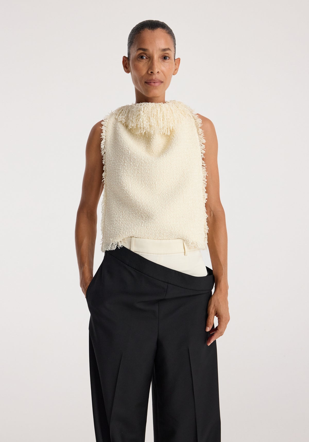 Woman wearing a sleeveless textured fringe bouclé top cream with a high neckline and frayed edges, paired with high-waisted black wide-leg pants.
