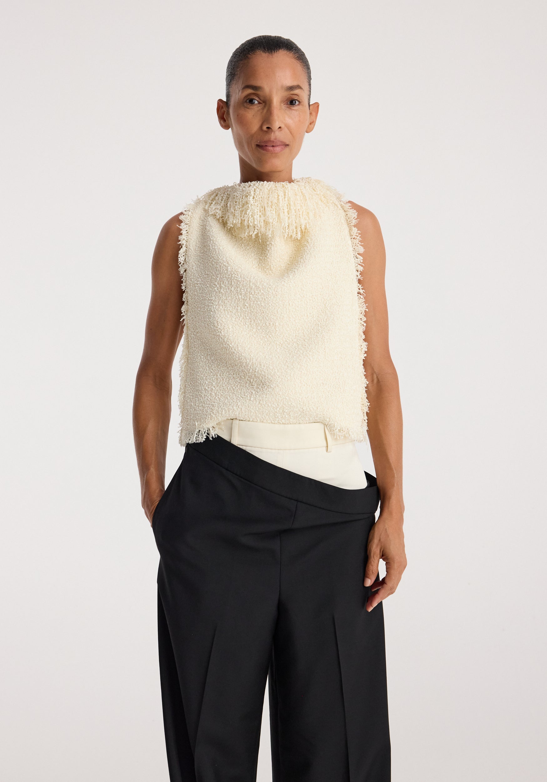 Woman wearing a sleeveless textured fringe bouclé top cream with a high neckline and frayed edges, paired with high-waisted black wide-leg pants.