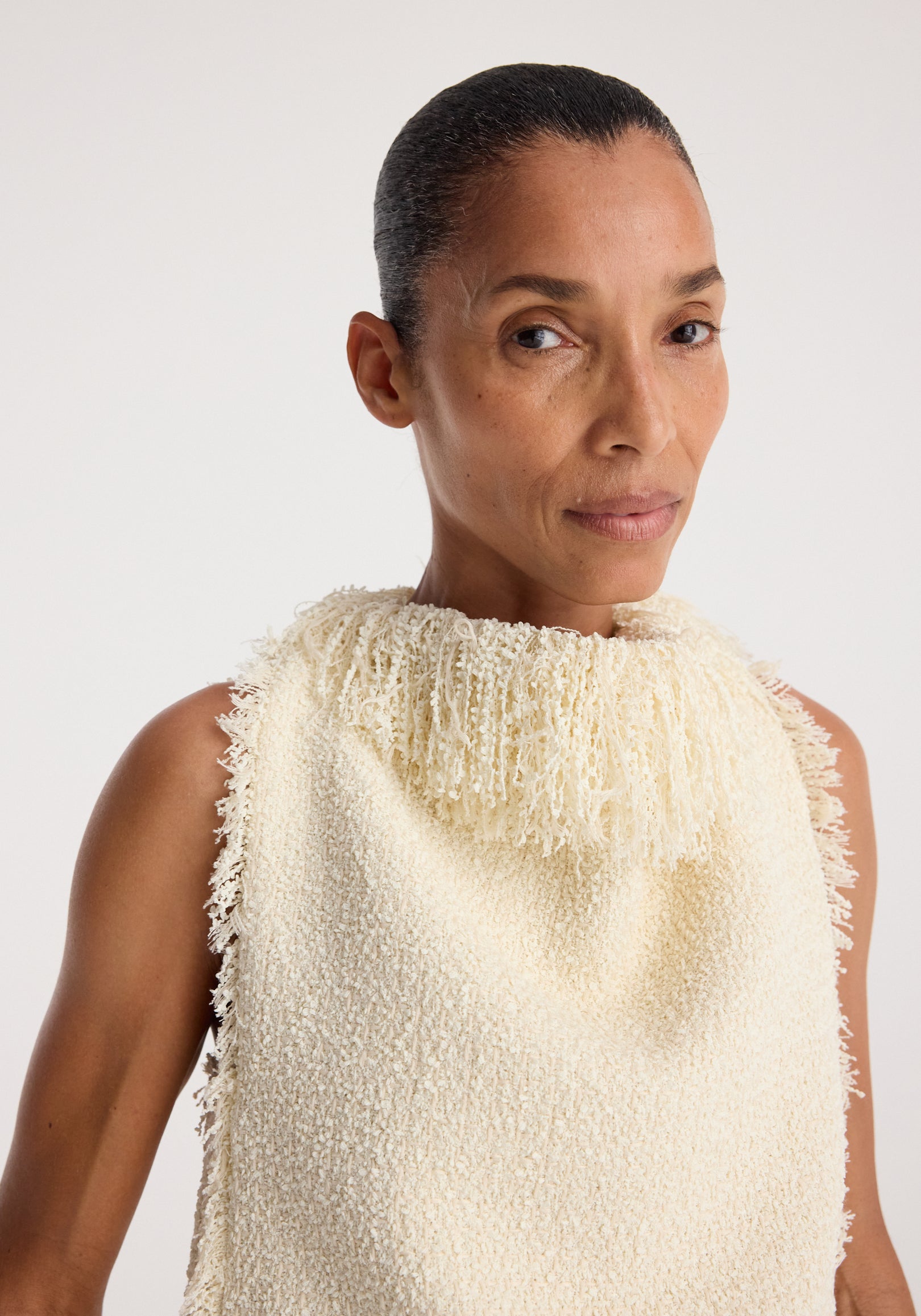 Close-up of a woman wearing a textured fringe bouclé top | cream with a high neckline and fringe detailing along the collar and edges against a white background.