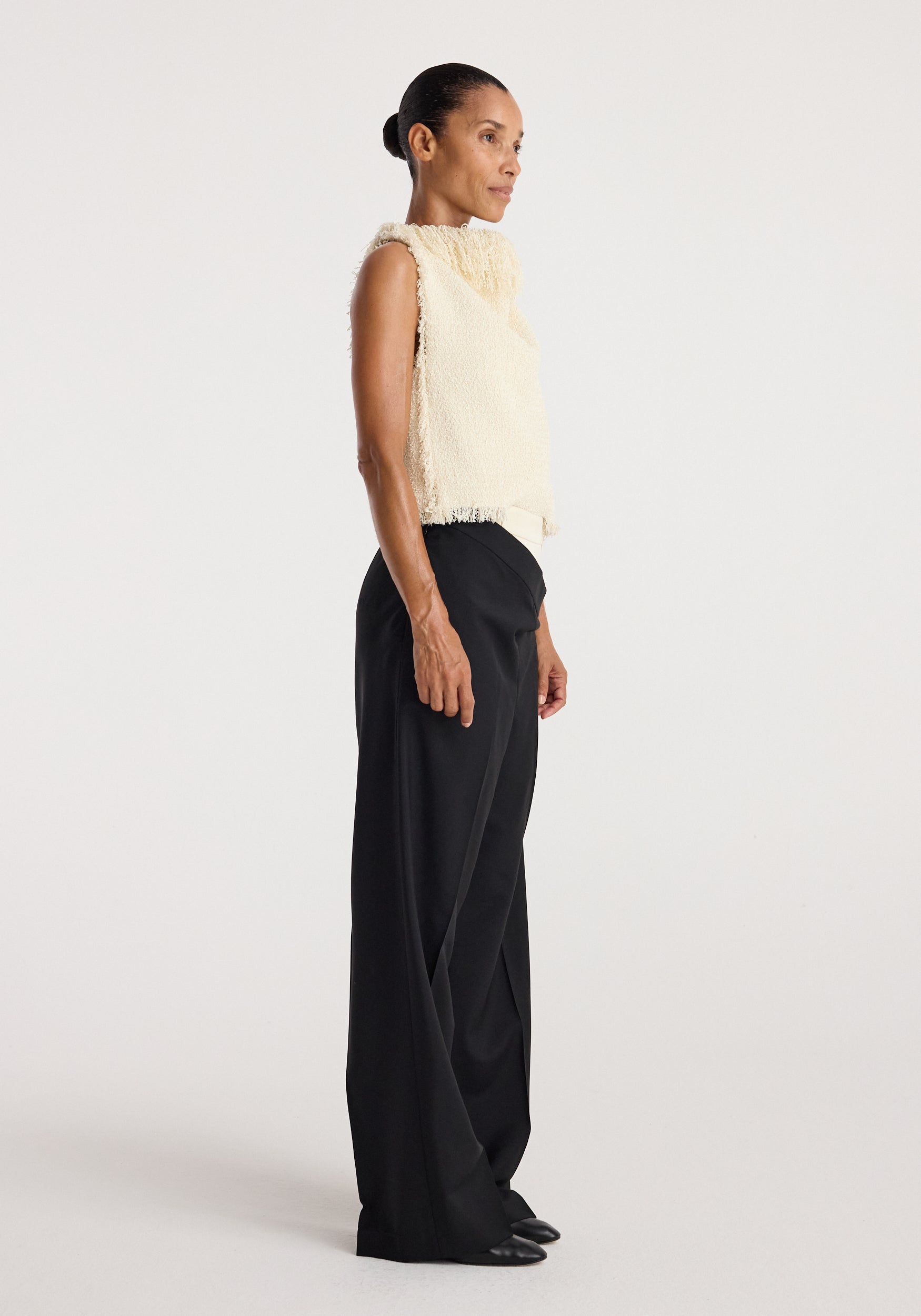 Side view of a woman wearing a sleeveless textured fringe bouclé top in cream with high-neck detail, paired with loose black pants and black shoes against a plain background.