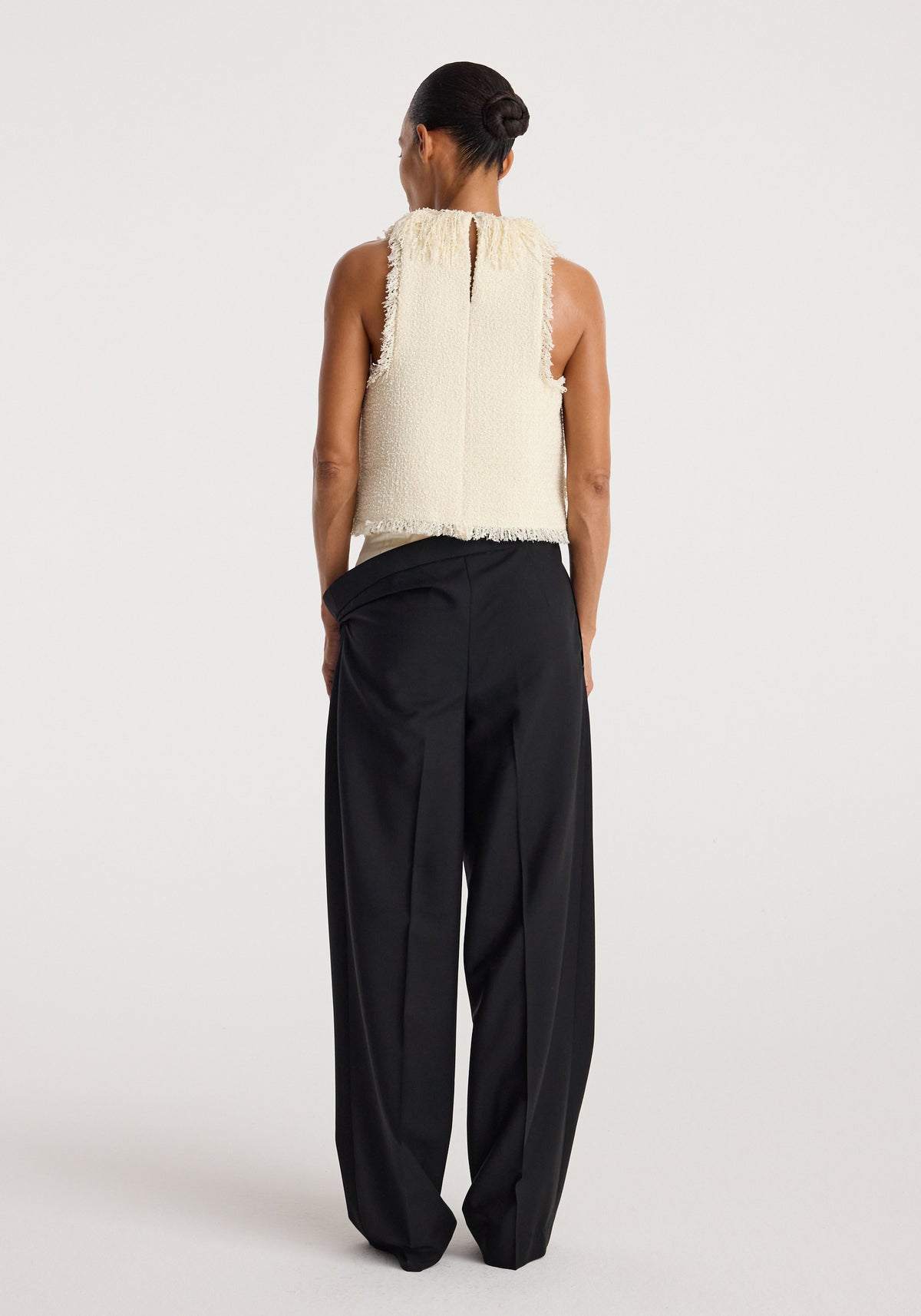 Back view of a woman wearing a sleeveless textured fringe bouclé top | cream paired with loose black trousers against a plain light background.