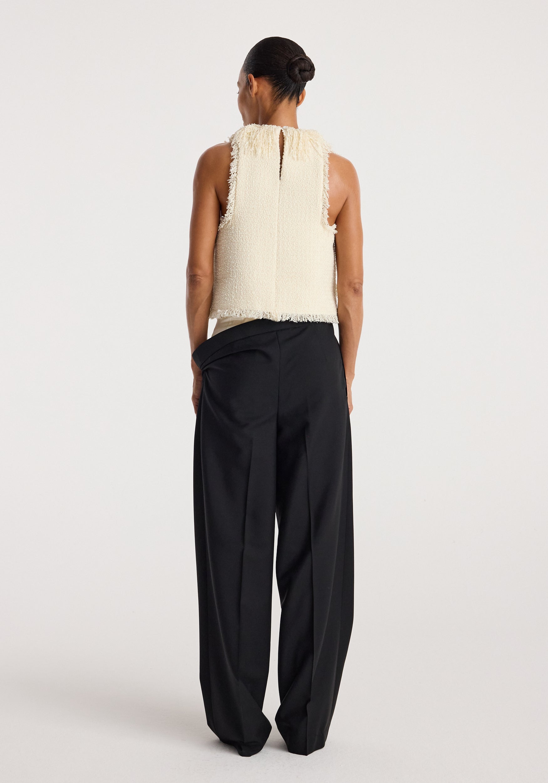 Back view of a woman wearing a sleeveless textured fringe bouclé top | cream paired with loose black trousers against a plain light background.