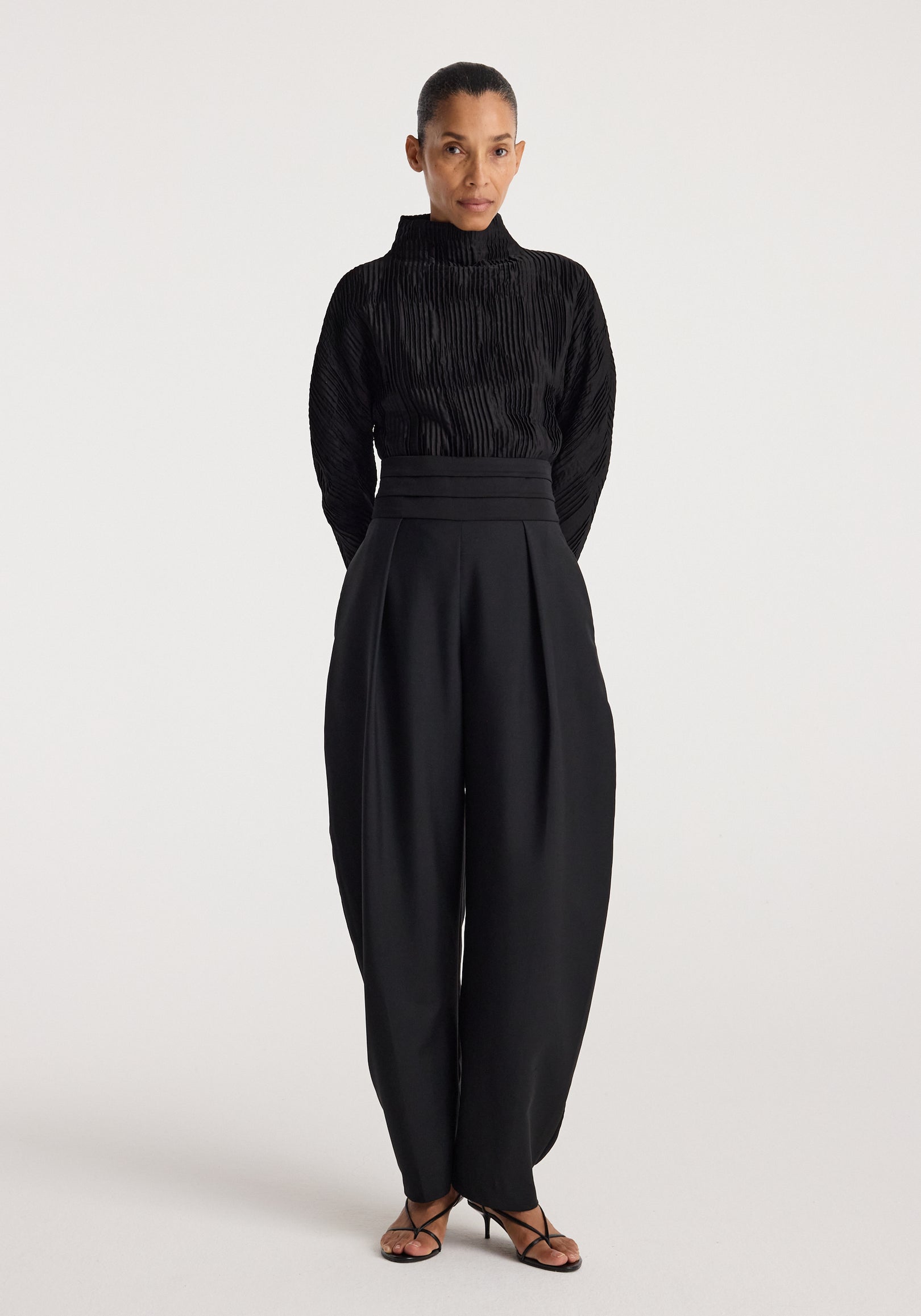 Woman wearing a black Plissé shaped top | black with long sleeves and high collar, paired with high-waisted wide-leg black pants and black strappy heels, standing against a plain background.