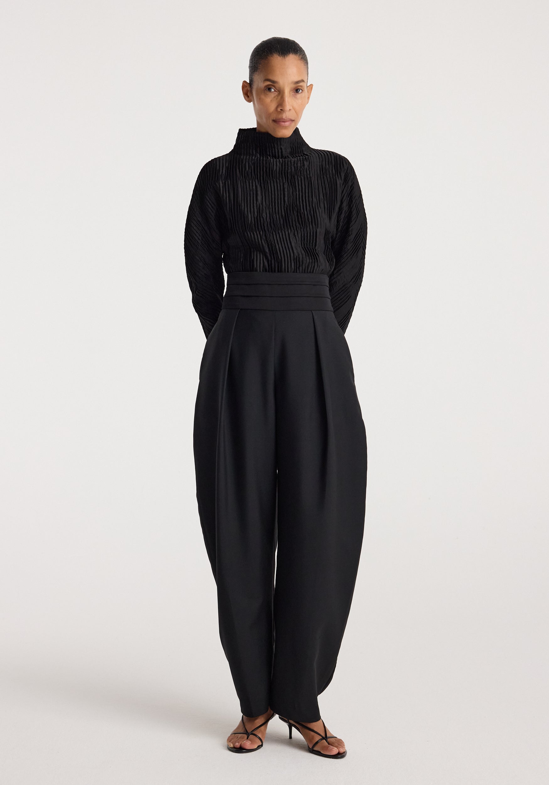 Woman wearing a black Plissé shaped top | black with long sleeves and high collar, paired with high-waisted wide-leg black pants and black strappy heels, standing against a plain background.