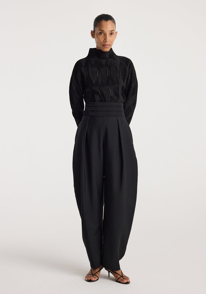 Woman wearing a black Plissé shaped top | black with long sleeves and high collar, paired with high-waisted wide-leg black pants and black strappy heels, standing against a plain background.