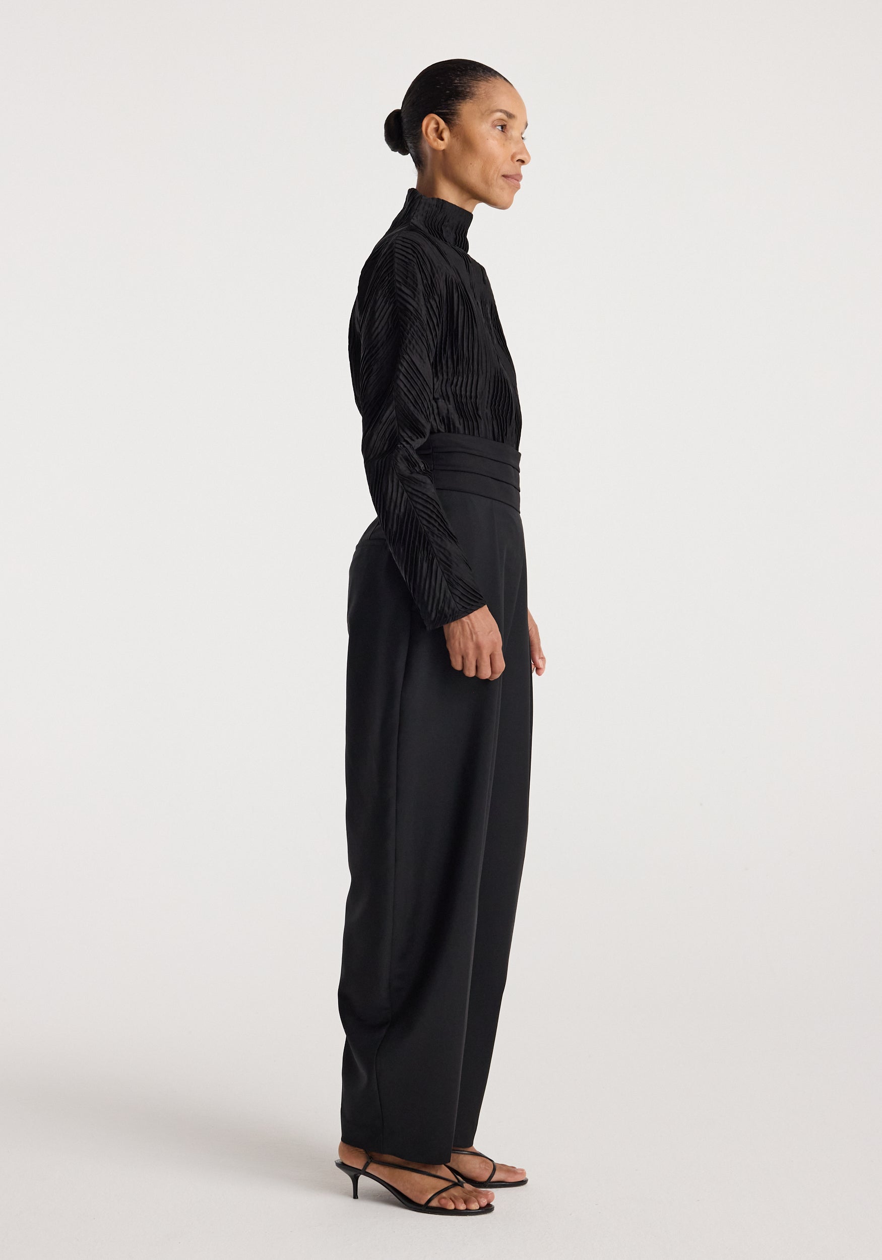 Woman wearing a black Plissé shaped top | black with long sleeves and a high neck, paired with high-waisted black trousers and black heeled sandals, standing in profile.