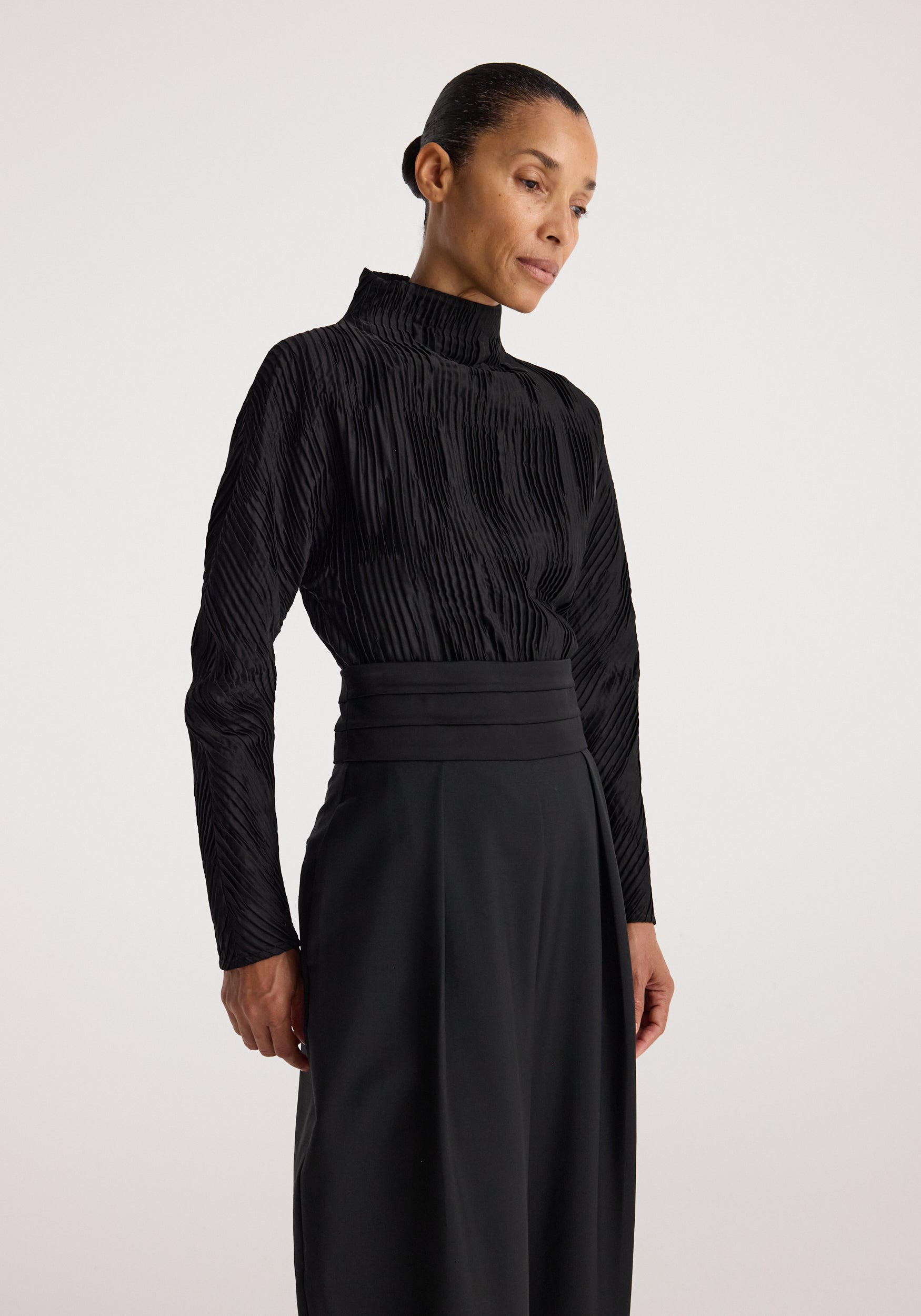 Woman wearing a black Plissé shaped top | black with long sleeves and a high neckline, paired with a high-waisted black skirt against a plain background.