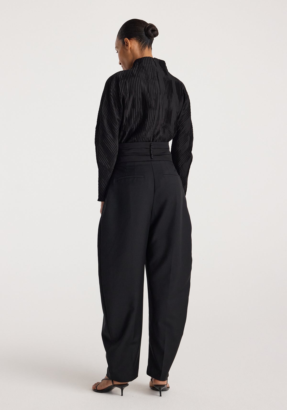 Woman wearing a black Plissé shaped top | black with long sleeves and textured pleats, shown from the back paired with high-waisted black pants and heeled sandals.