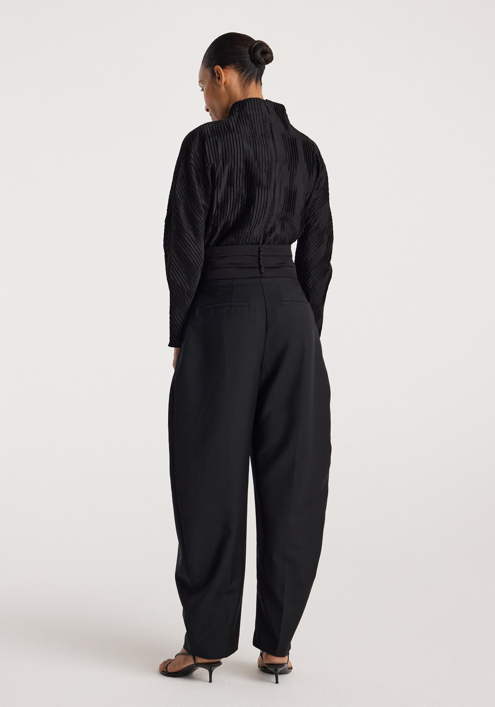 Woman wearing a black Plissé shaped top | black with long sleeves and textured pleats, shown from the back paired with high-waisted black pants and heeled sandals.