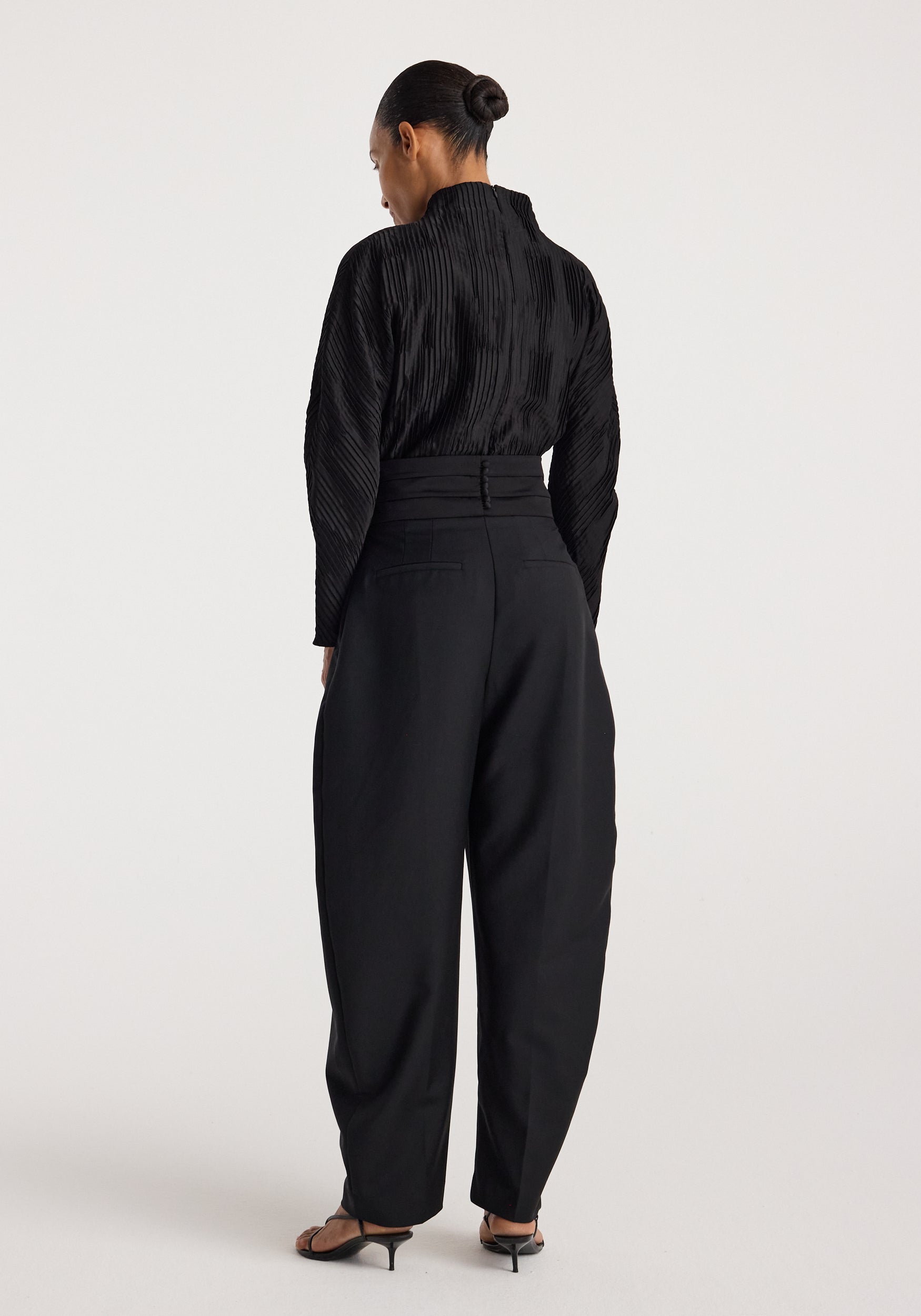 Woman wearing a black Plissé shaped top | black with long sleeves and textured pleats, shown from the back paired with high-waisted black pants and heeled sandals.