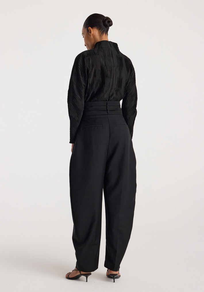 Woman wearing a black Plissé shaped top | black with long sleeves and textured pleats, shown from the back paired with high-waisted black pants and heeled sandals.