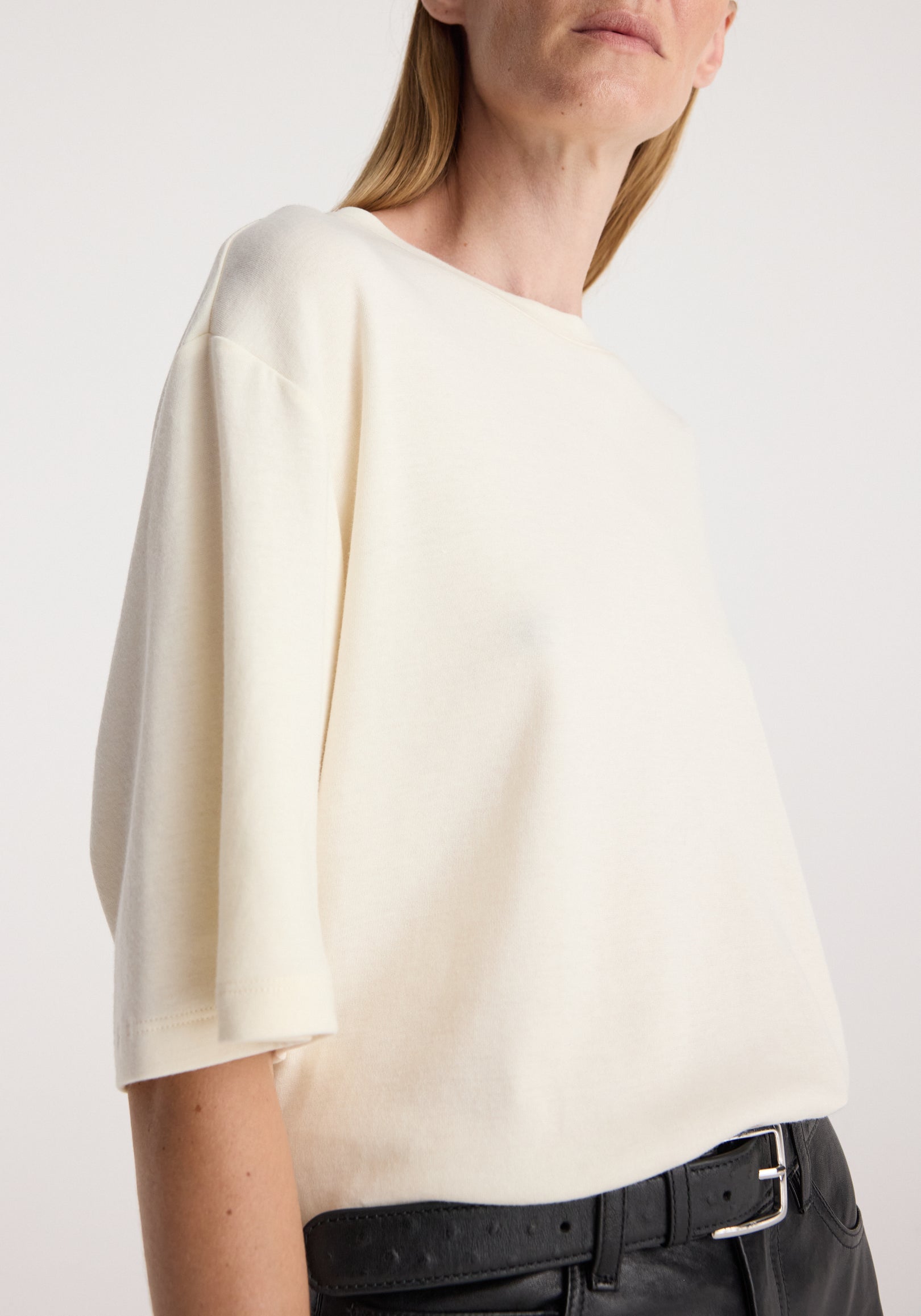 Woman wearing an oversized cotton wool t-shirt | cream with a round neckline and half sleeves, paired with black belt and pants against a plain background.
