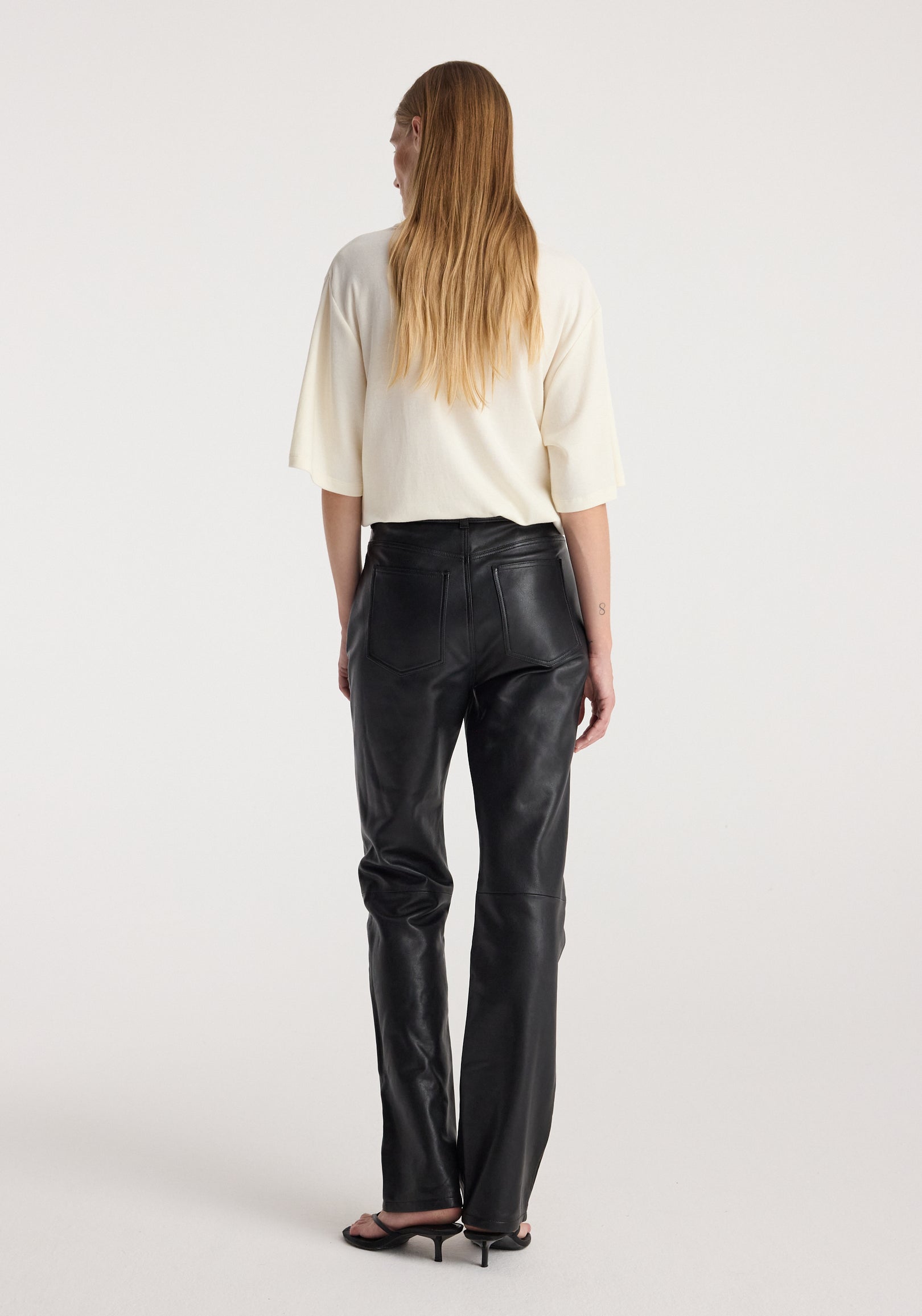 Woman seen from behind wearing an oversized cotton wool t-shirt | cream paired with black leather pants and black heeled sandals against a plain background.