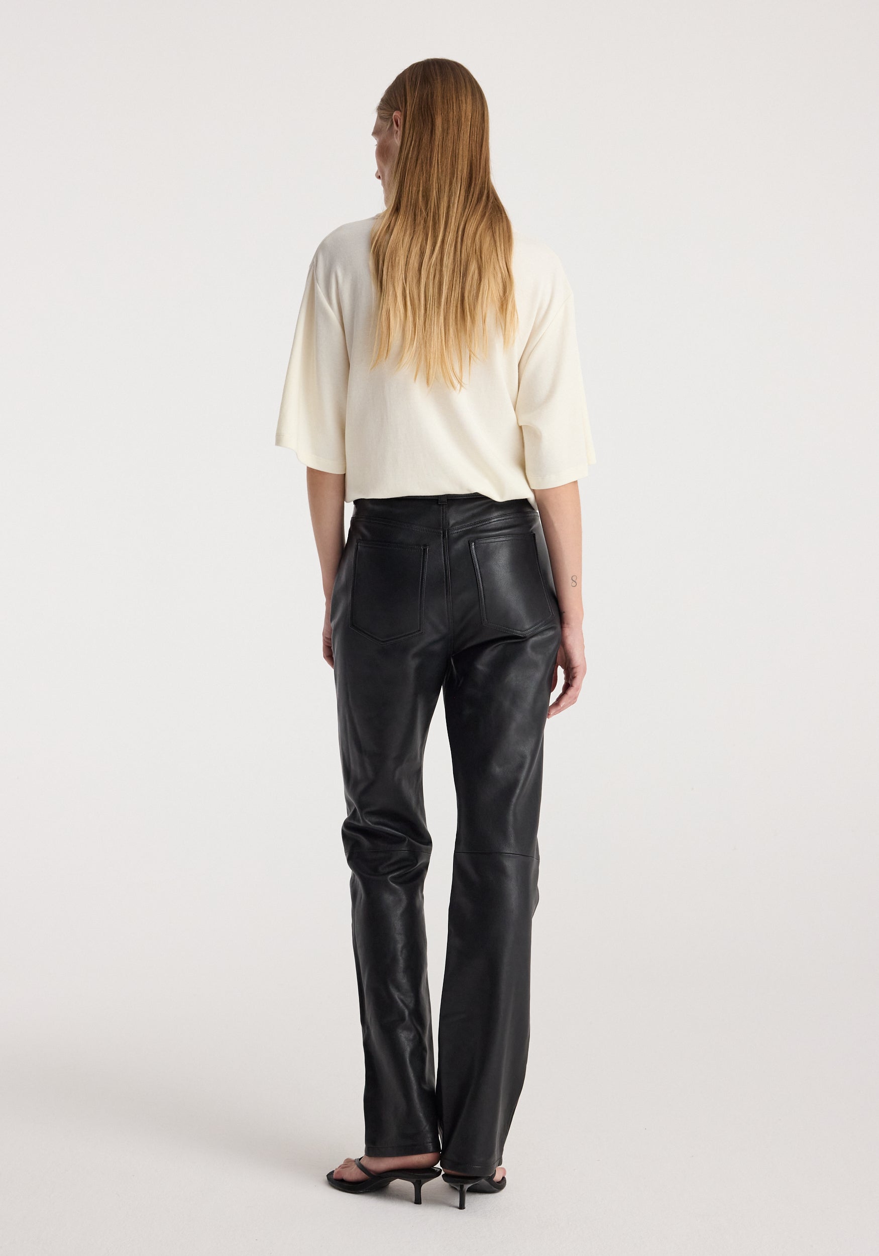 Woman seen from behind wearing an oversized cotton wool t-shirt | cream paired with black leather pants and black heeled sandals against a plain background.
