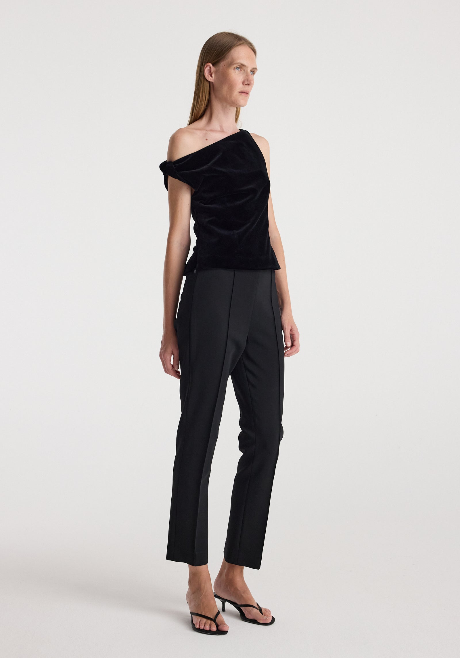 Woman wearing a draped velvet top | black with one-shoulder design paired with black tailored pants and black heeled sandals against a plain background.