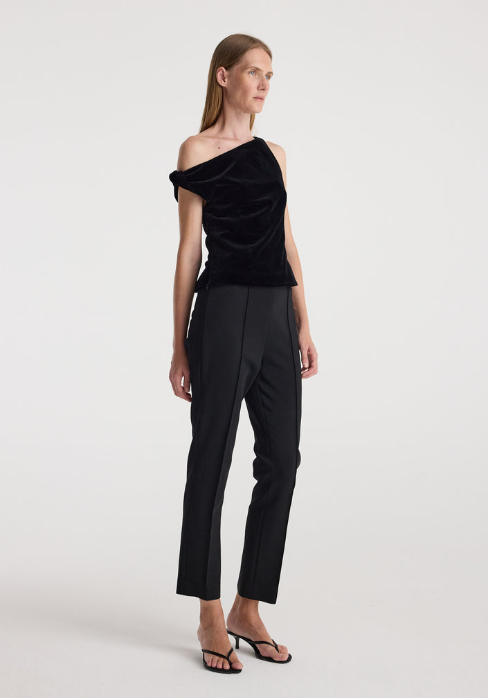 Woman wearing a draped velvet top | black with one-shoulder design paired with black tailored pants and black heeled sandals against a plain background.
