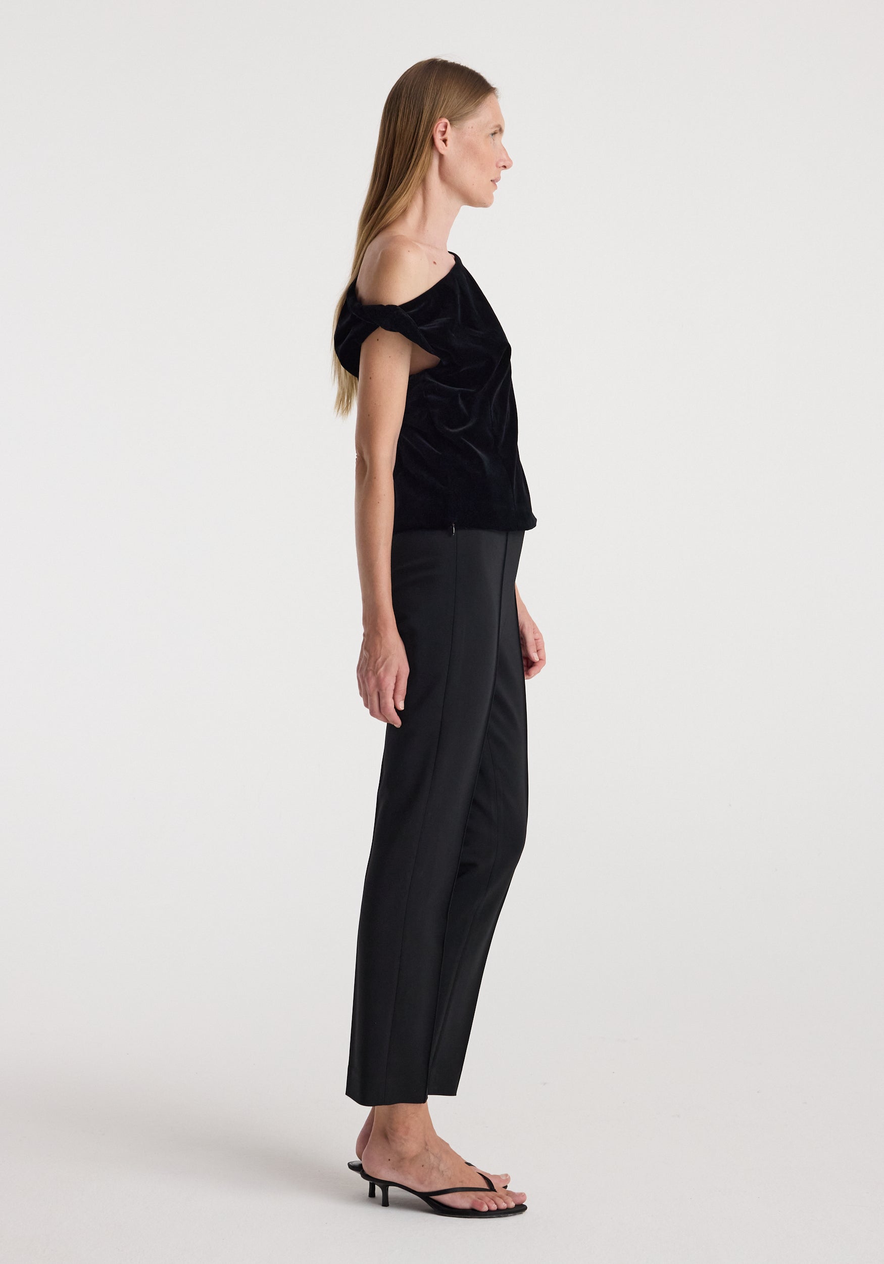 Woman wearing a black draped velvet top | black with an off-the-shoulder design paired with black tailored pants and black open-toe heeled sandals against a white background.