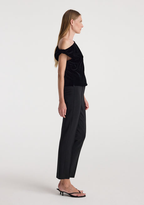 Woman wearing a draped velvet top | black with one-shoulder design paired with black tailored pants and black heeled sandals against a plain background.