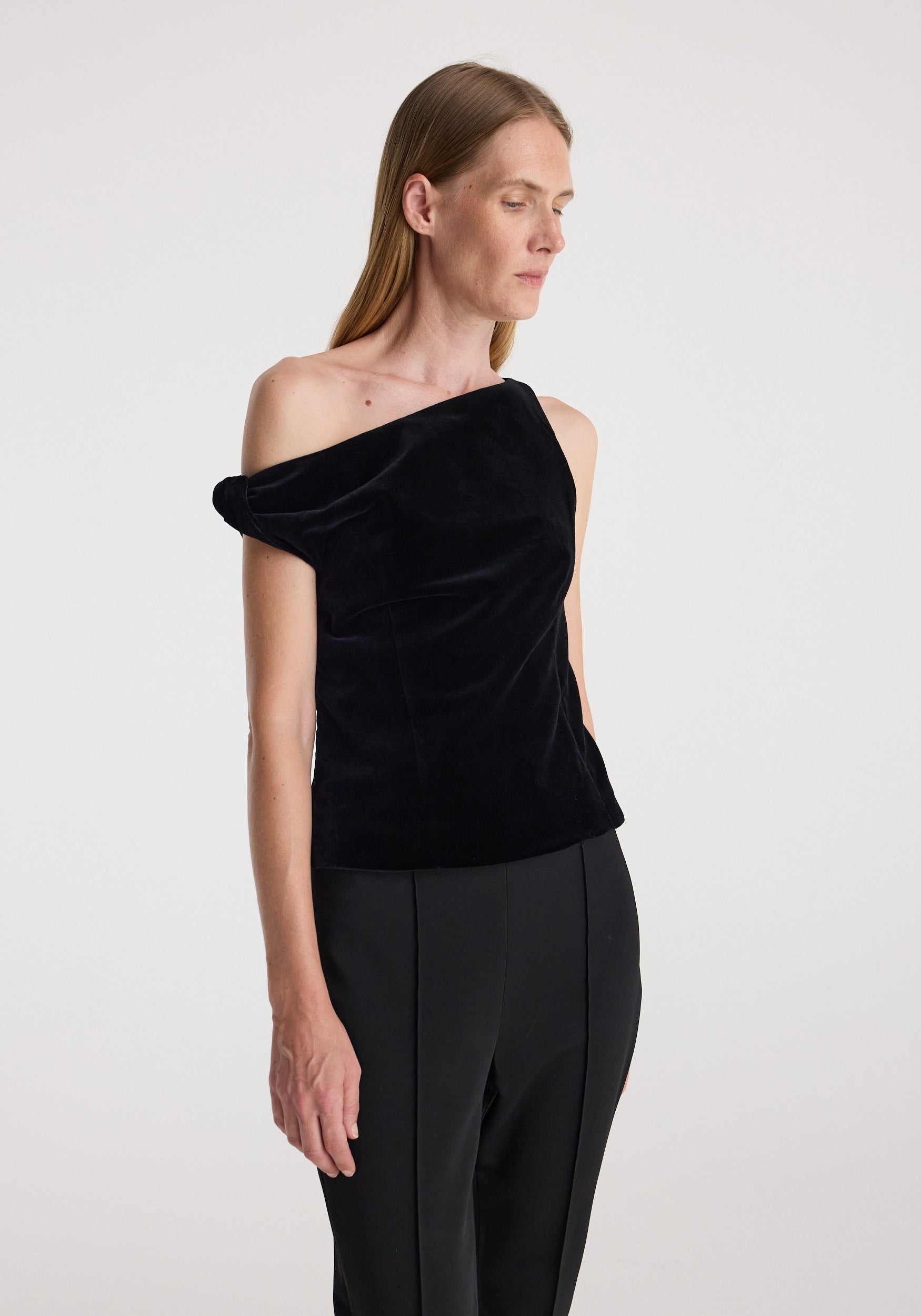 Woman wearing a black draped velvet top | black with an asymmetrical off-shoulder design paired with black trousers against a plain background.