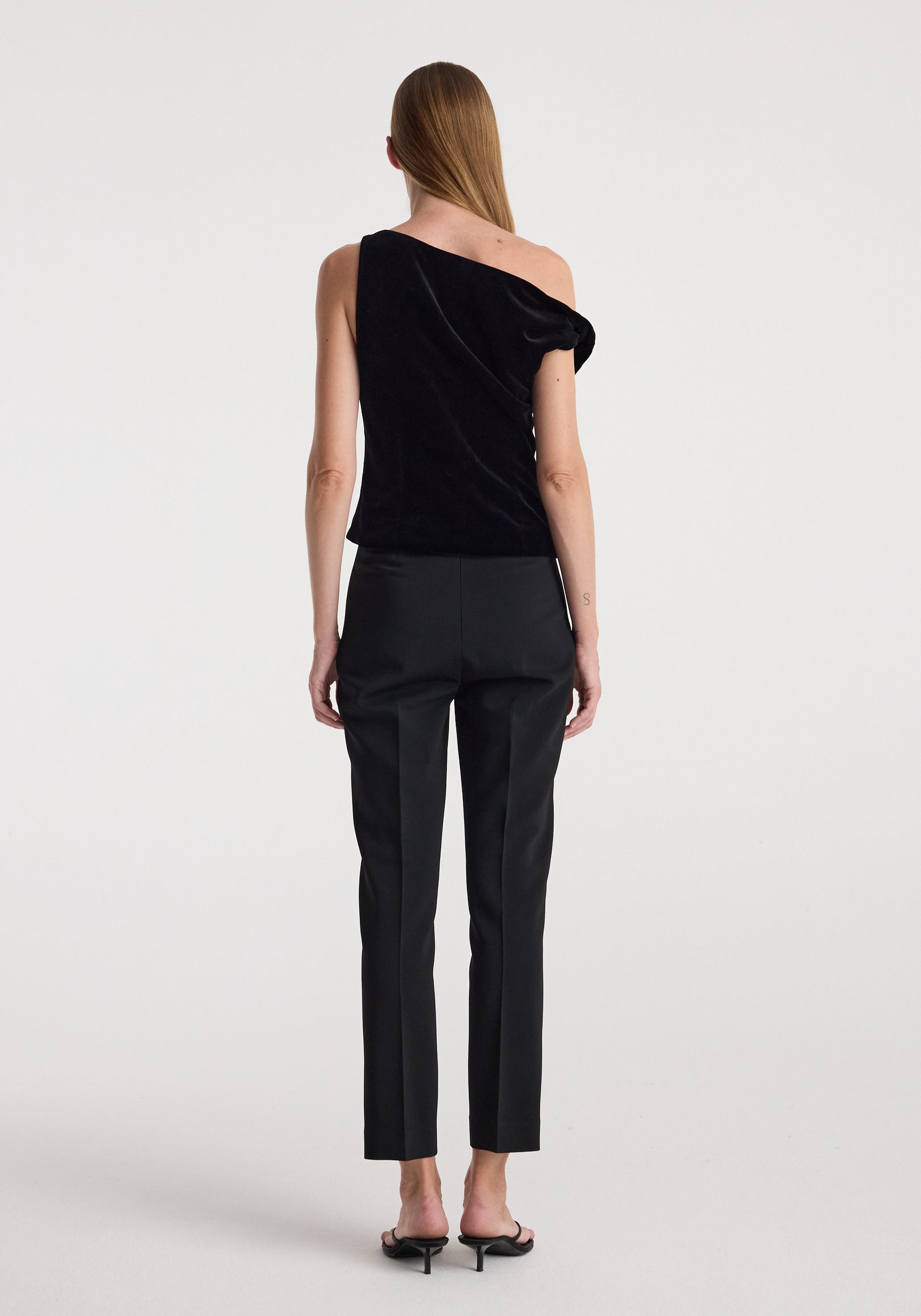 Woman wearing a black Draped velvet top | black with one shoulder exposed, paired with black tailored pants and black heeled sandals, standing against a plain white background.