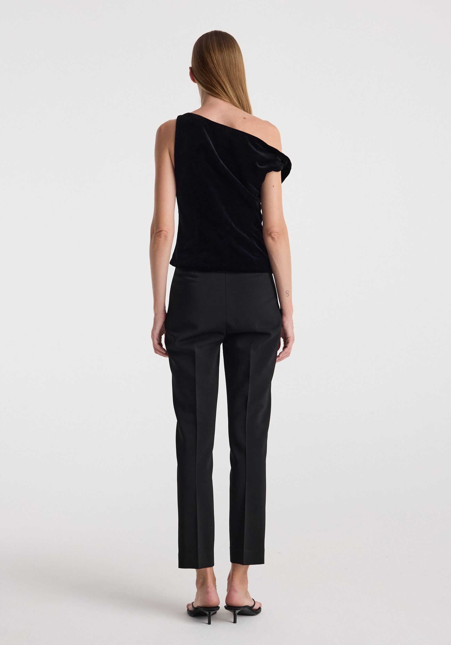 Woman wearing a black Draped velvet top | black with one shoulder exposed, paired with black tailored pants and black heeled sandals, standing against a plain white background.