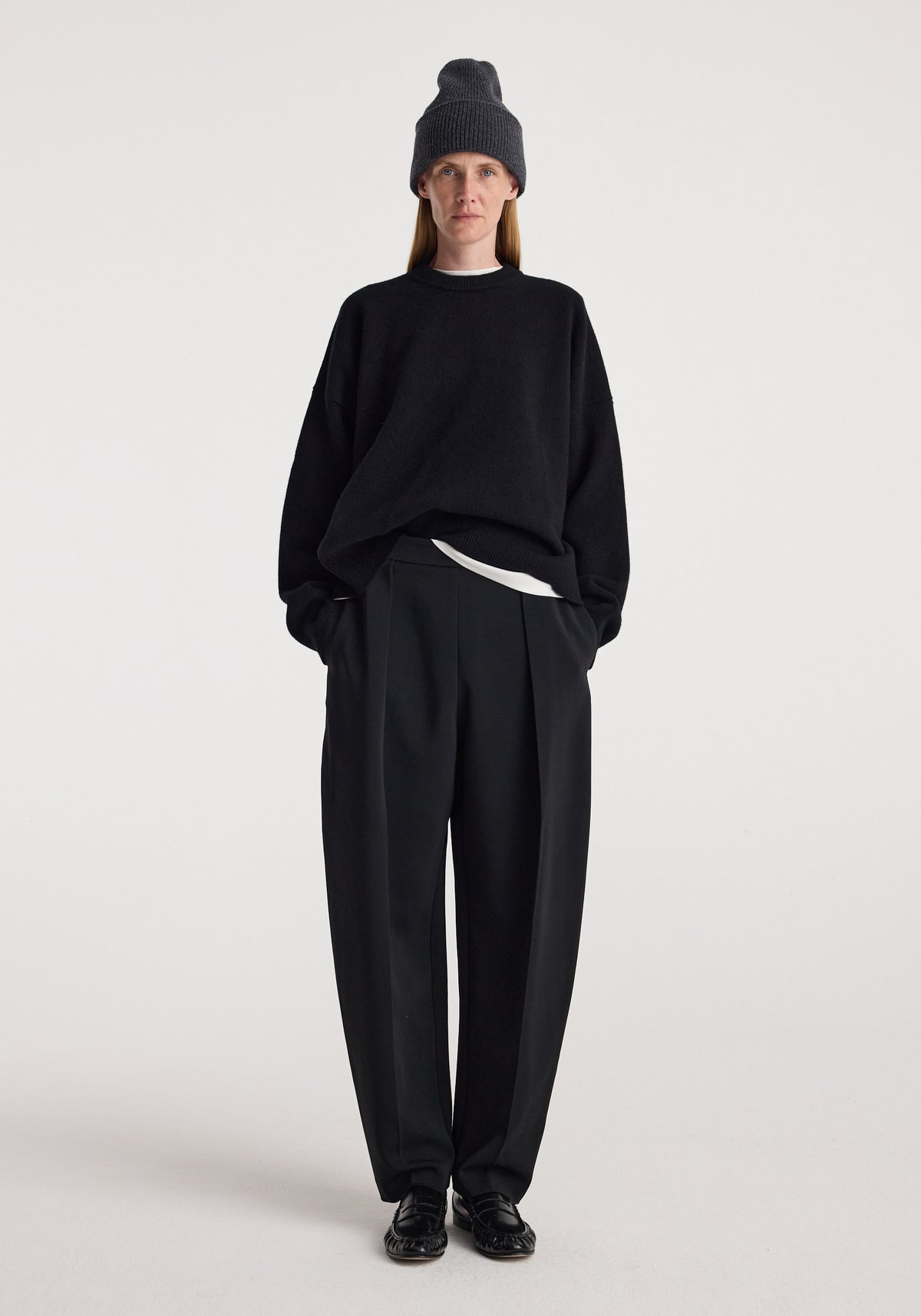 Person wearing a Beanie | anthracite melange styled with a black oversized sweater, black pleated trousers, and black shoes against a plain white background.