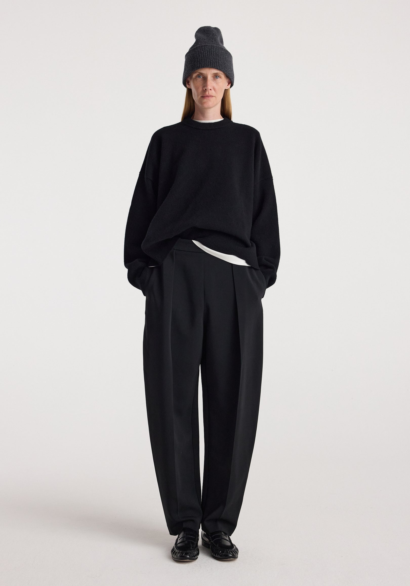 Person wearing a Beanie | anthracite melange styled with a black oversized sweater, black pleated trousers, and black shoes against a plain white background.