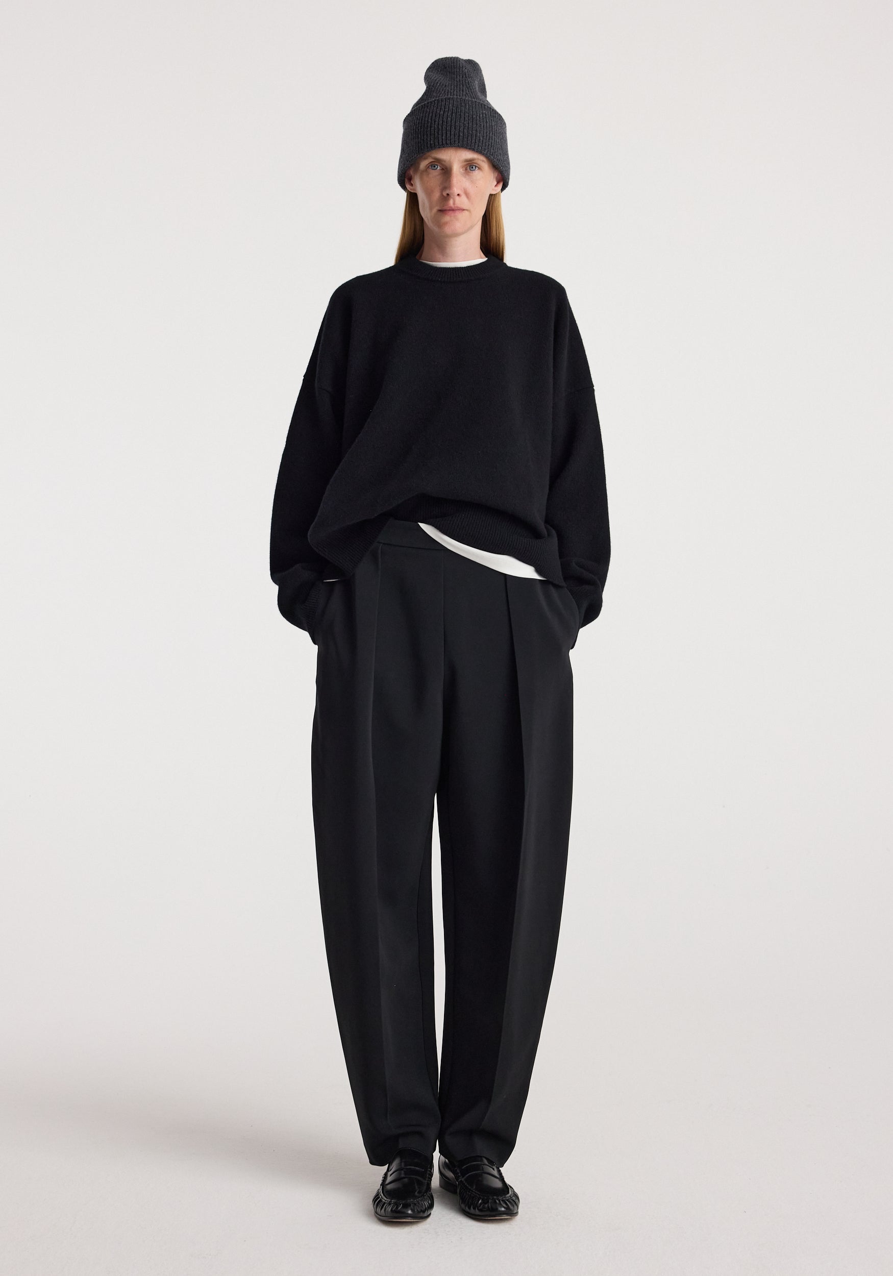 Person wearing a Beanie | anthracite melange styled with a black oversized sweater, black pleated trousers, and black shoes against a plain white background.