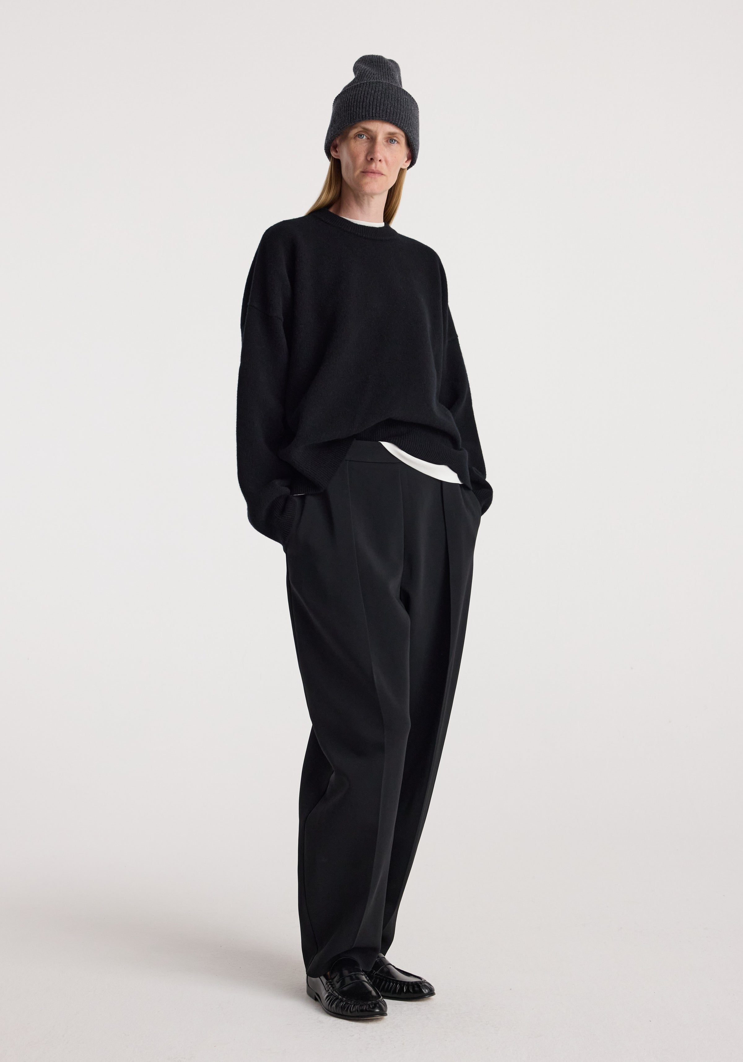 Person wearing a dark grey knitted Beanie | anthracite melange with black long-sleeve sweater, black pants, and black shoes standing against a plain light background.