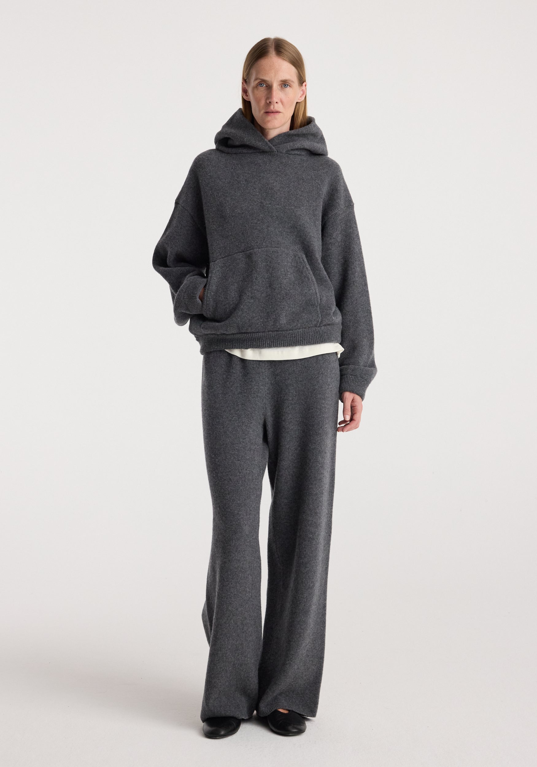 Woman wearing dark grey melange knit hoodie and Compact knitted trousers | dark grey melange, standing against a plain light background with black shoes.