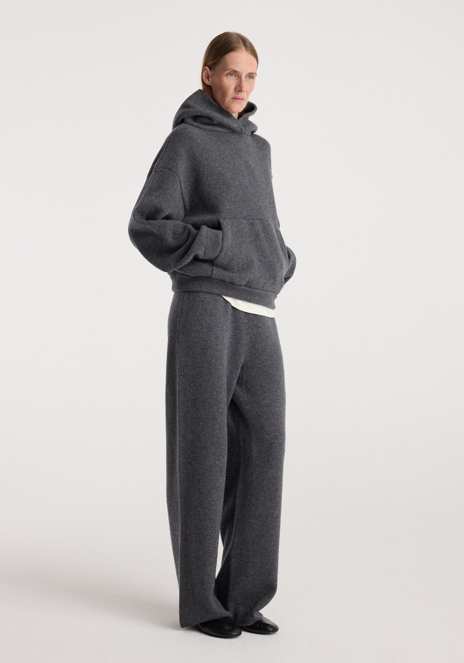 Woman wearing a dark grey melange knitted hoodie and Compact knitted trousers | dark grey melange, standing with hands in pockets against a plain light background.