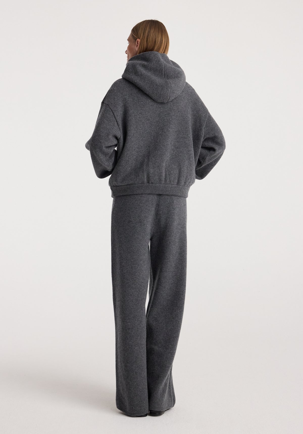 Woman wearing dark grey melange hooded sweater paired with Compact knitted trousers | dark grey melange standing against plain white background viewed from the back.