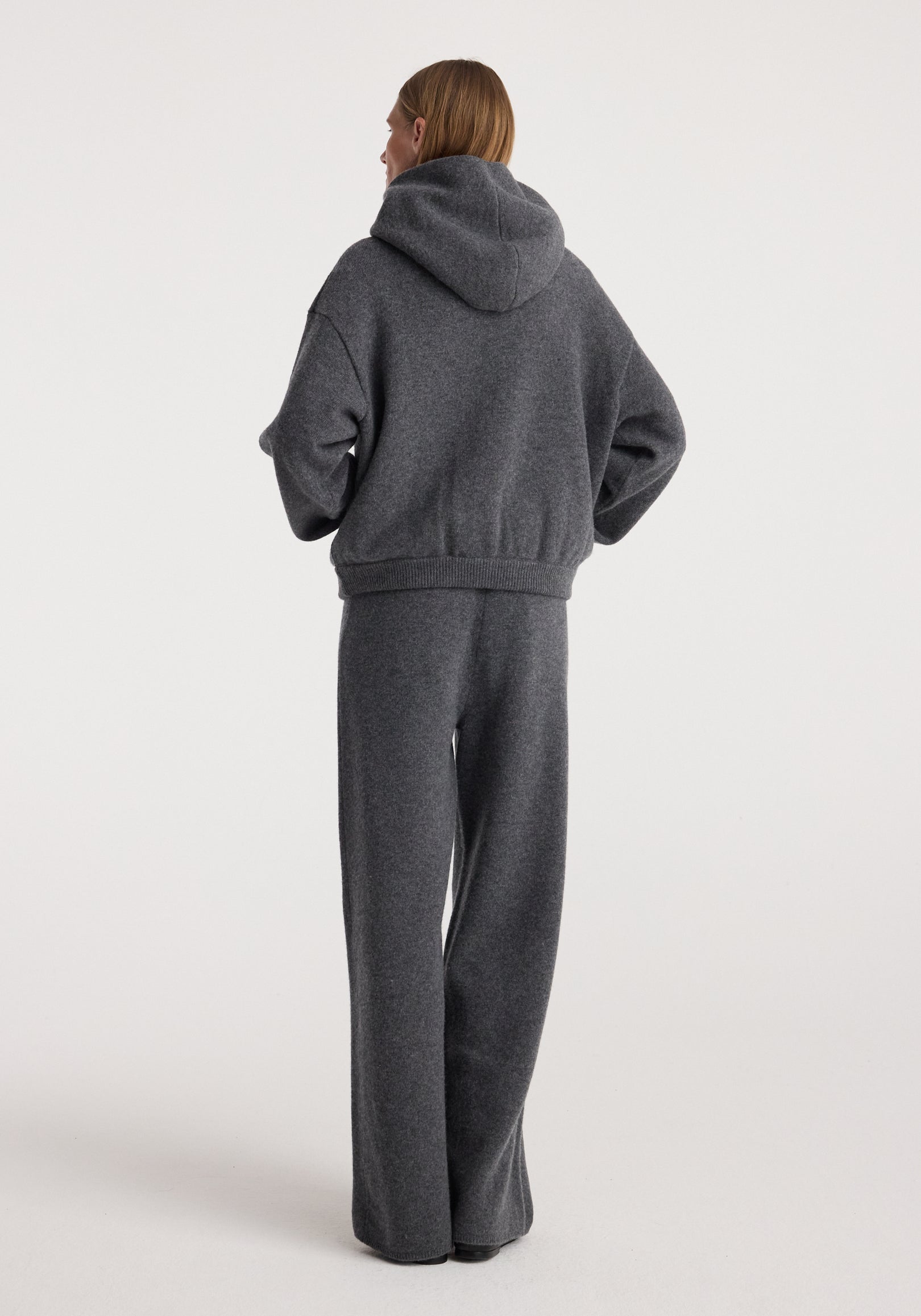 Woman wearing dark grey melange hooded sweater paired with Compact knitted trousers | dark grey melange standing against plain white background viewed from the back.