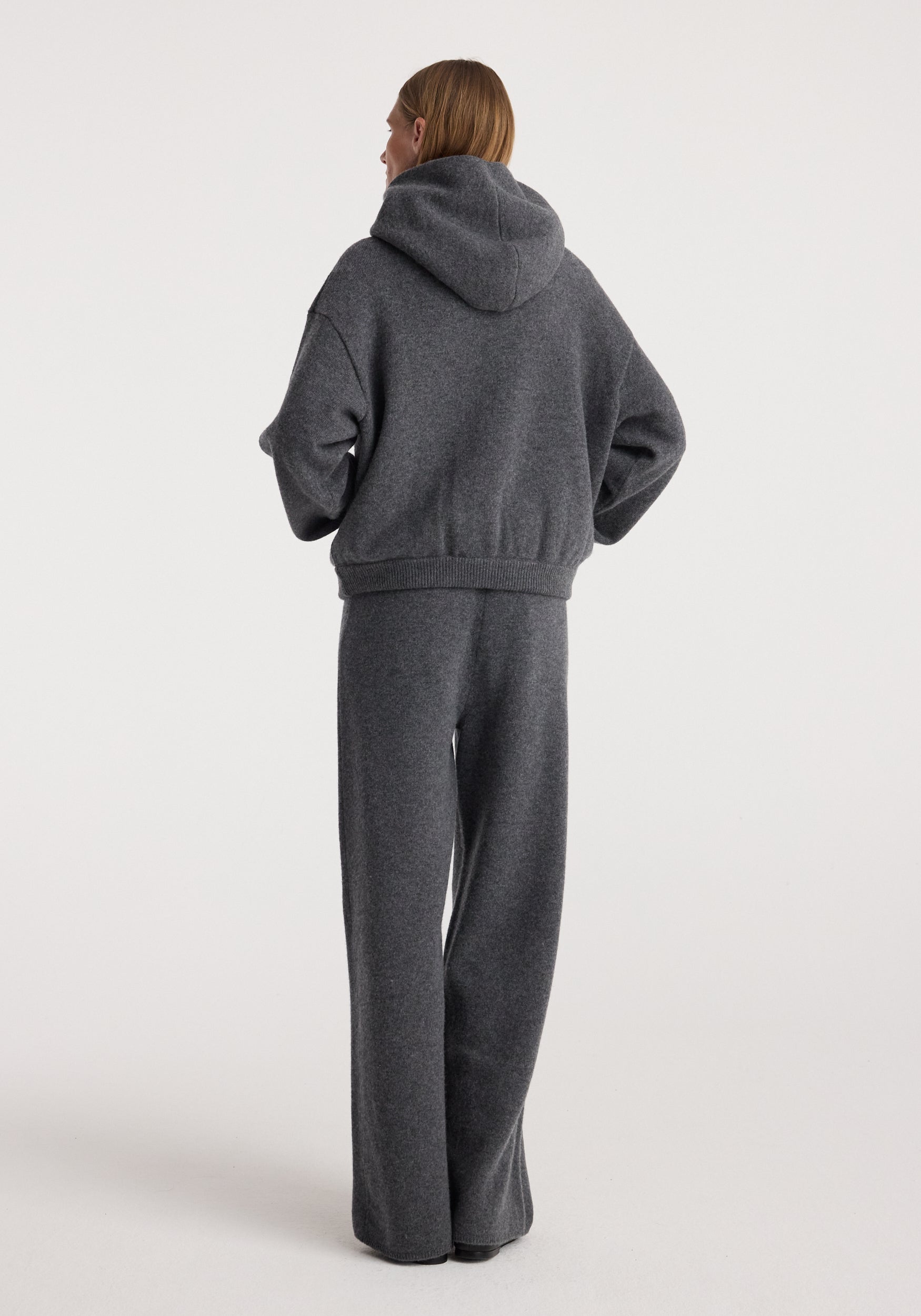 Woman wearing dark grey melange hooded sweater paired with Compact knitted trousers | dark grey melange standing against plain white background viewed from the back.