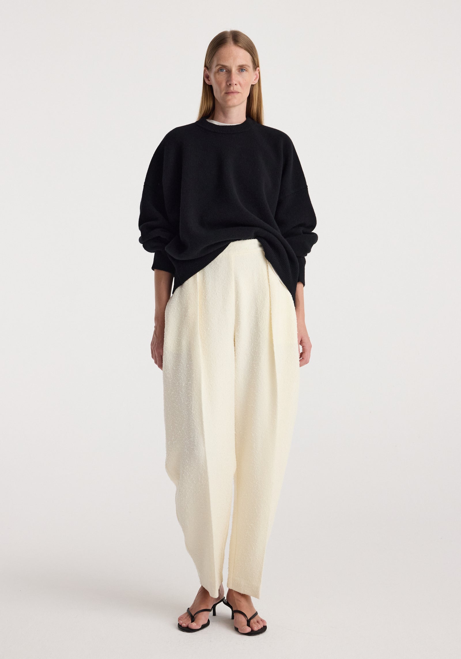 Woman wearing a black Classic crewneck jumper | black with loose white trousers and black open-toe heels against a plain light background.