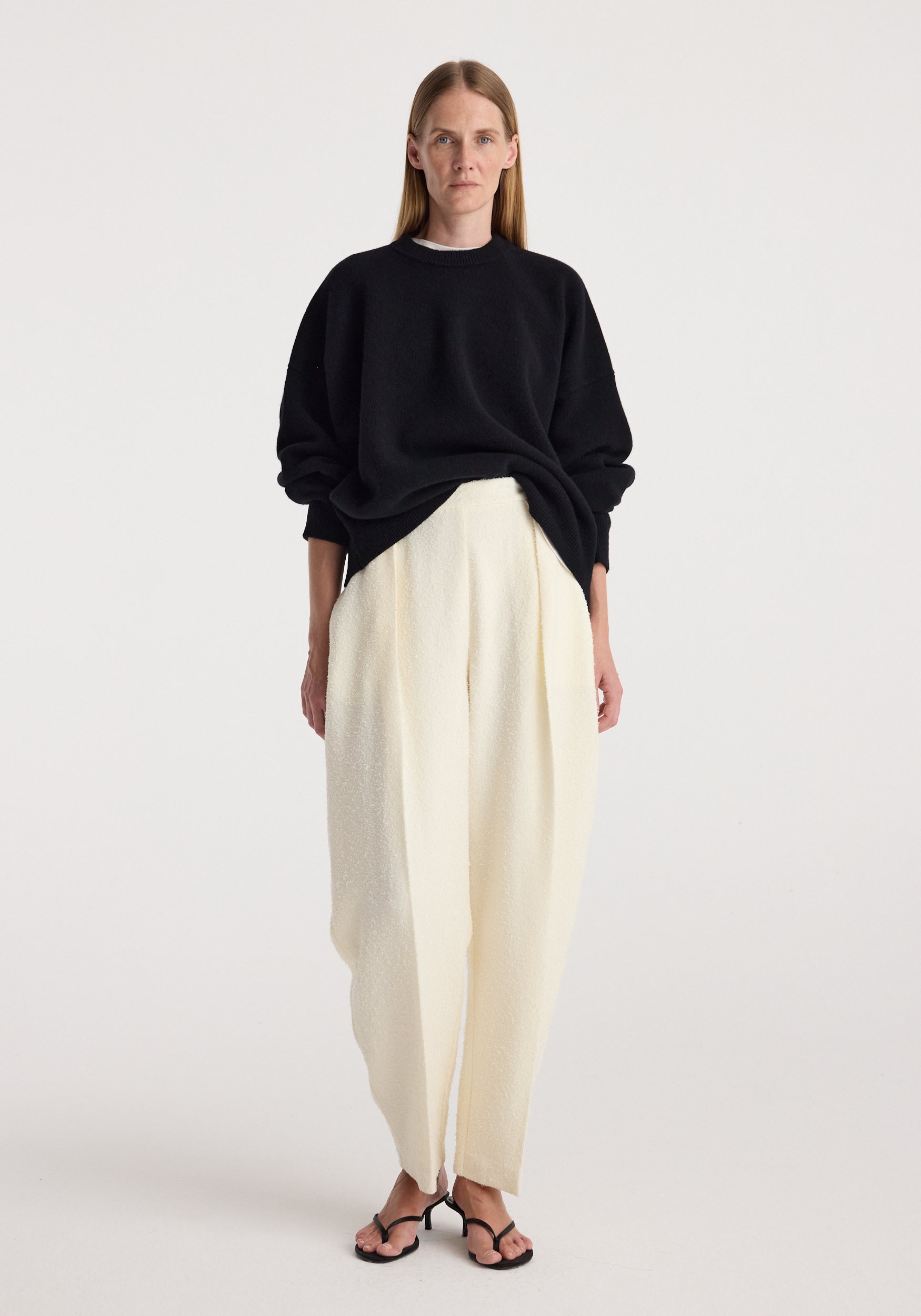 Woman wearing a black Classic crewneck jumper | black with loose white trousers and black open-toe heels against a plain light background.