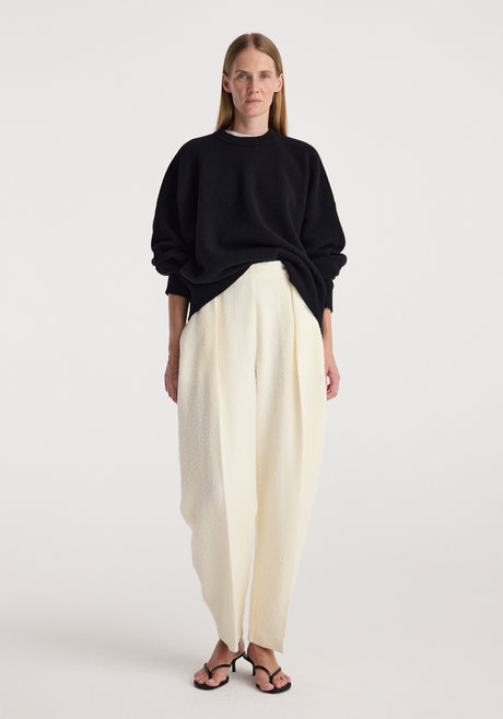 Woman wearing a black Classic crewneck jumper | black with loose white trousers and black open-toe heels against a plain light background.