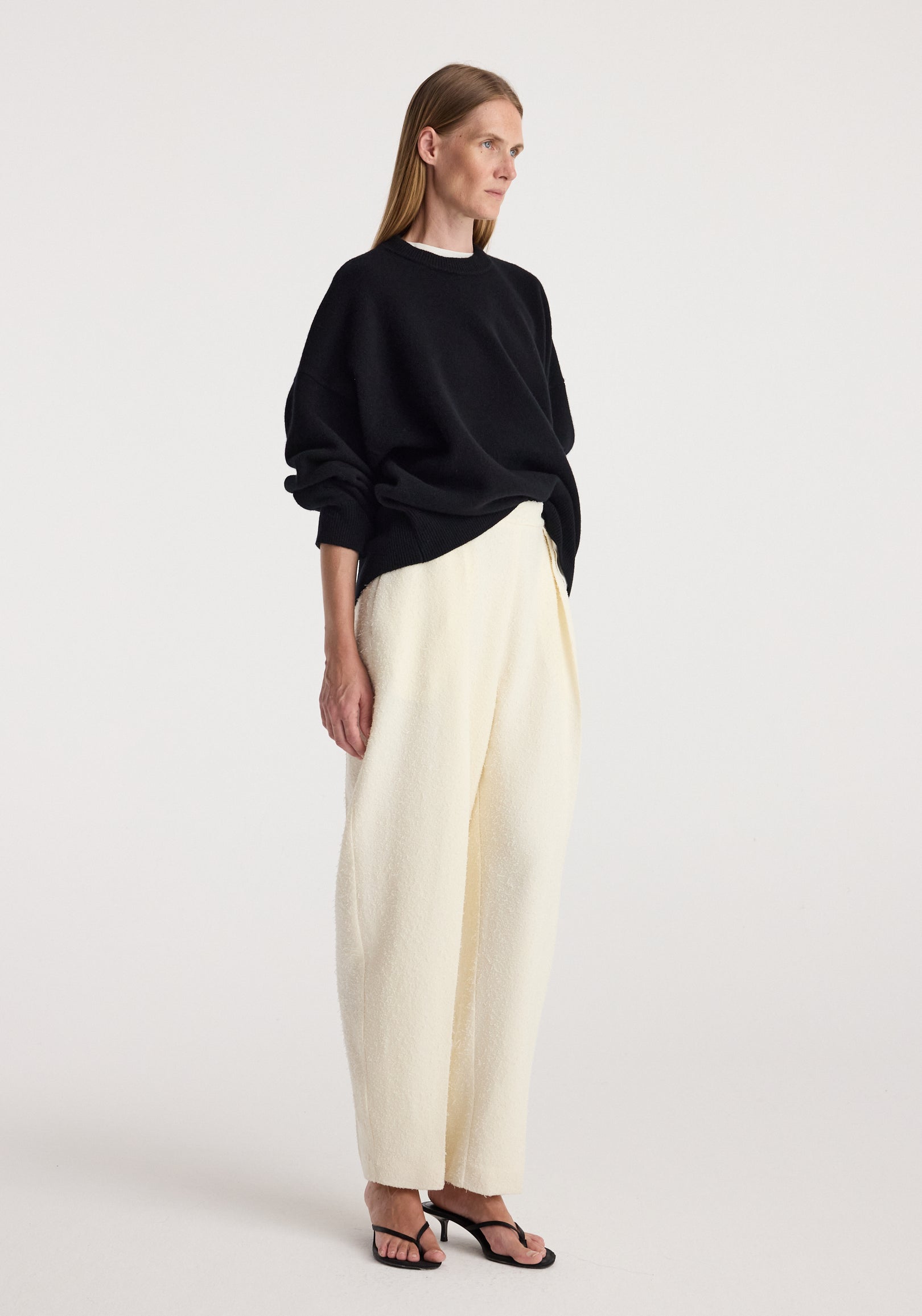Woman wearing a loose-fitting Classic crewneck jumper | black paired with cream wide-leg pants and black open-toe heeled sandals, standing against a plain background.